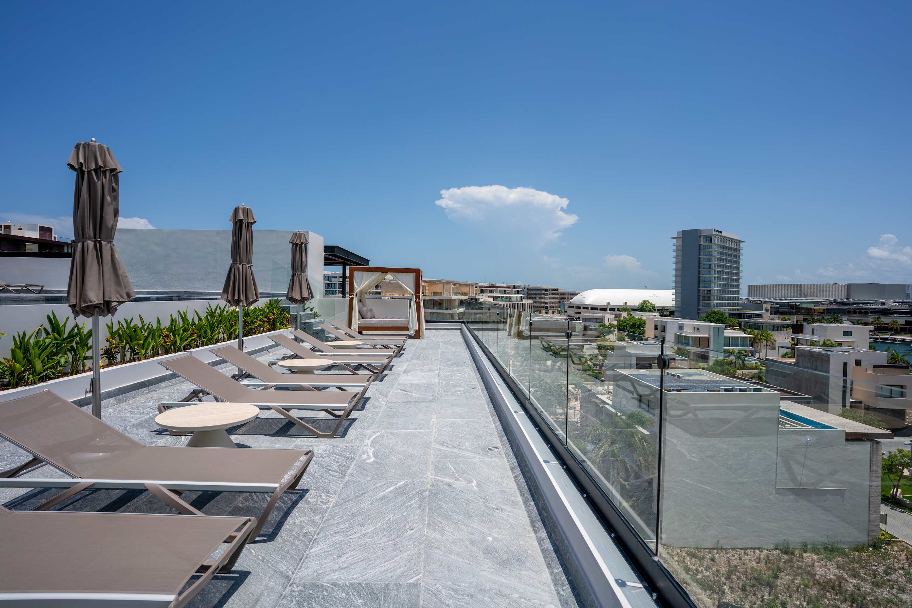 Rooftop lounge area with lounge chairs, umbrellas, and a canopy daybed, overlooking a cityscape.