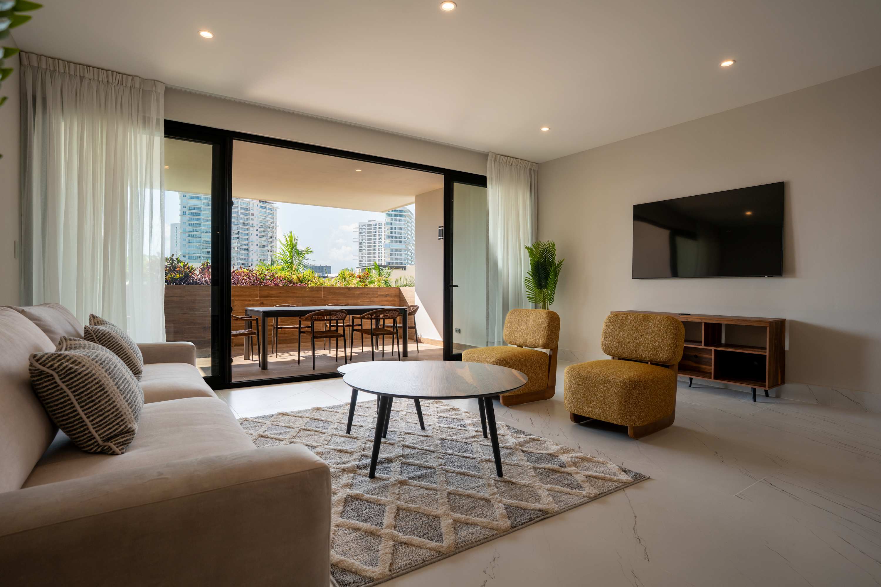 Living room with couch, chairs, and coffee table opens to a balcony with dining table and city view.