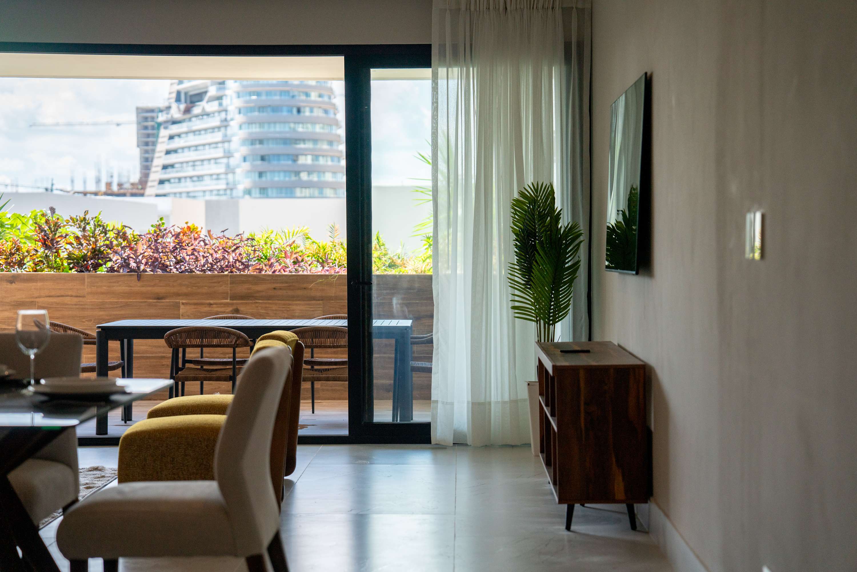 Modern living room with dining area and a view of a city and tropical plants.