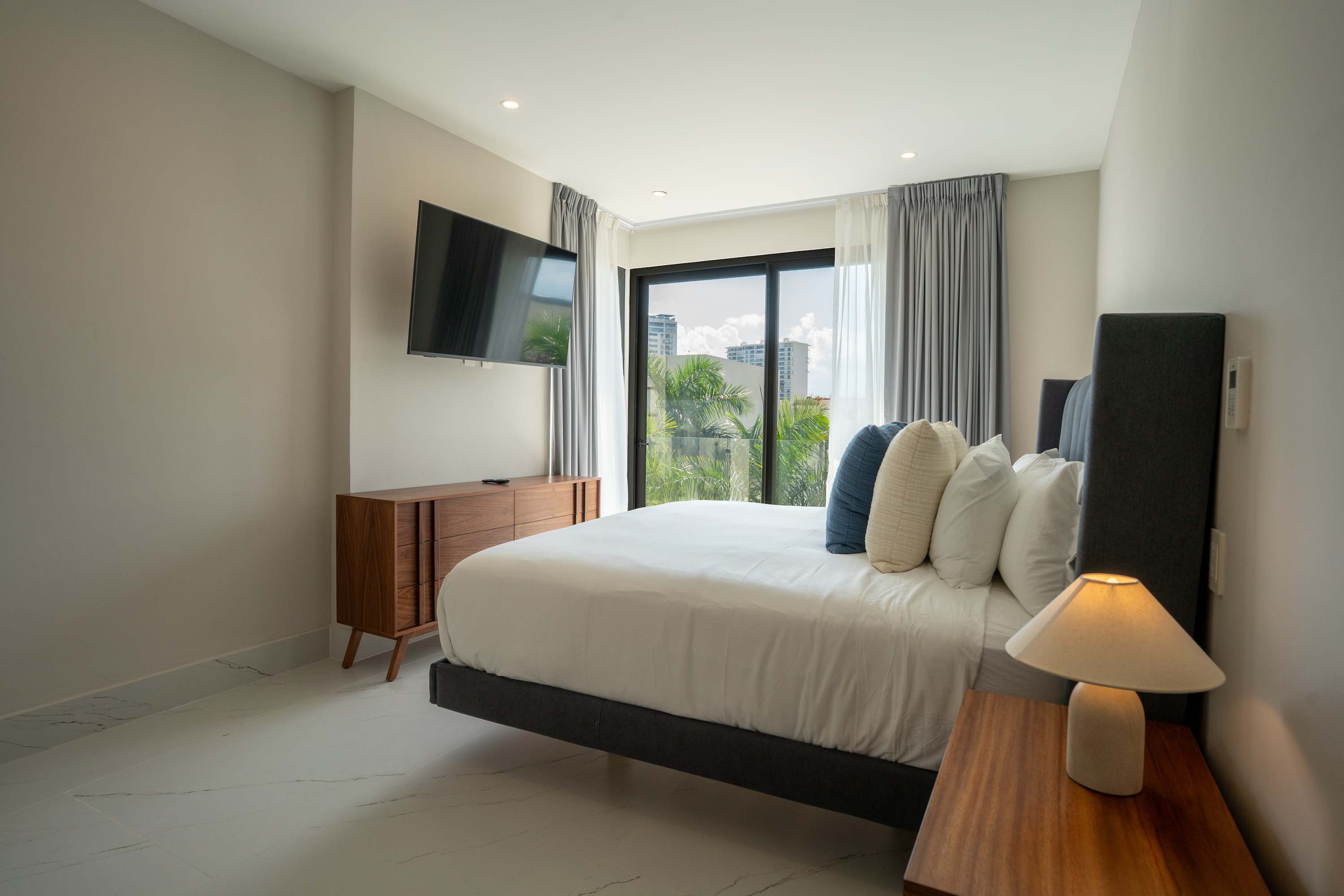 A modern bedroom with a large bed, TV, and a view of palm trees and buildings through a large window