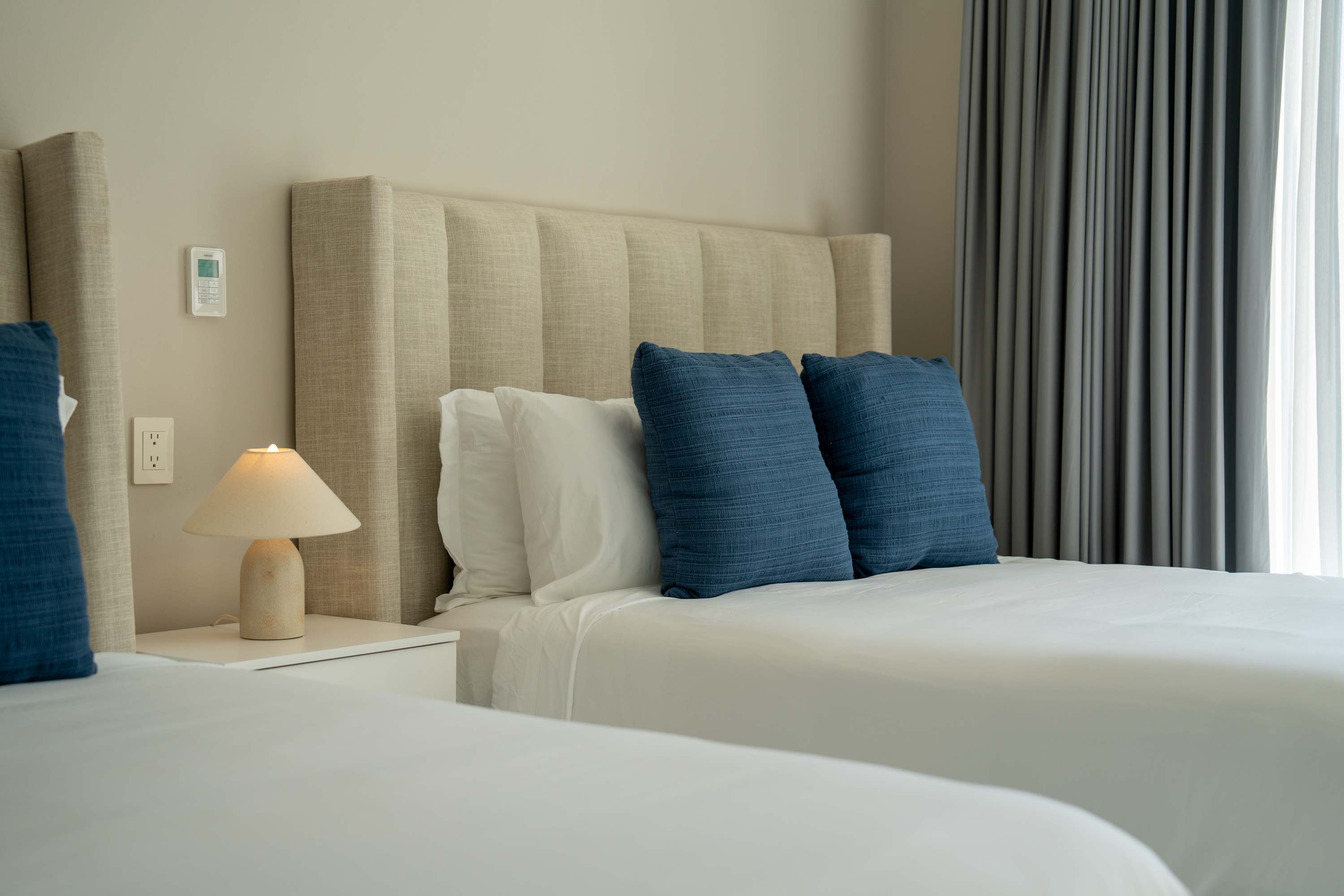 Two beds in a hotel room with white linens, blue pillows, and a bedside lamp.