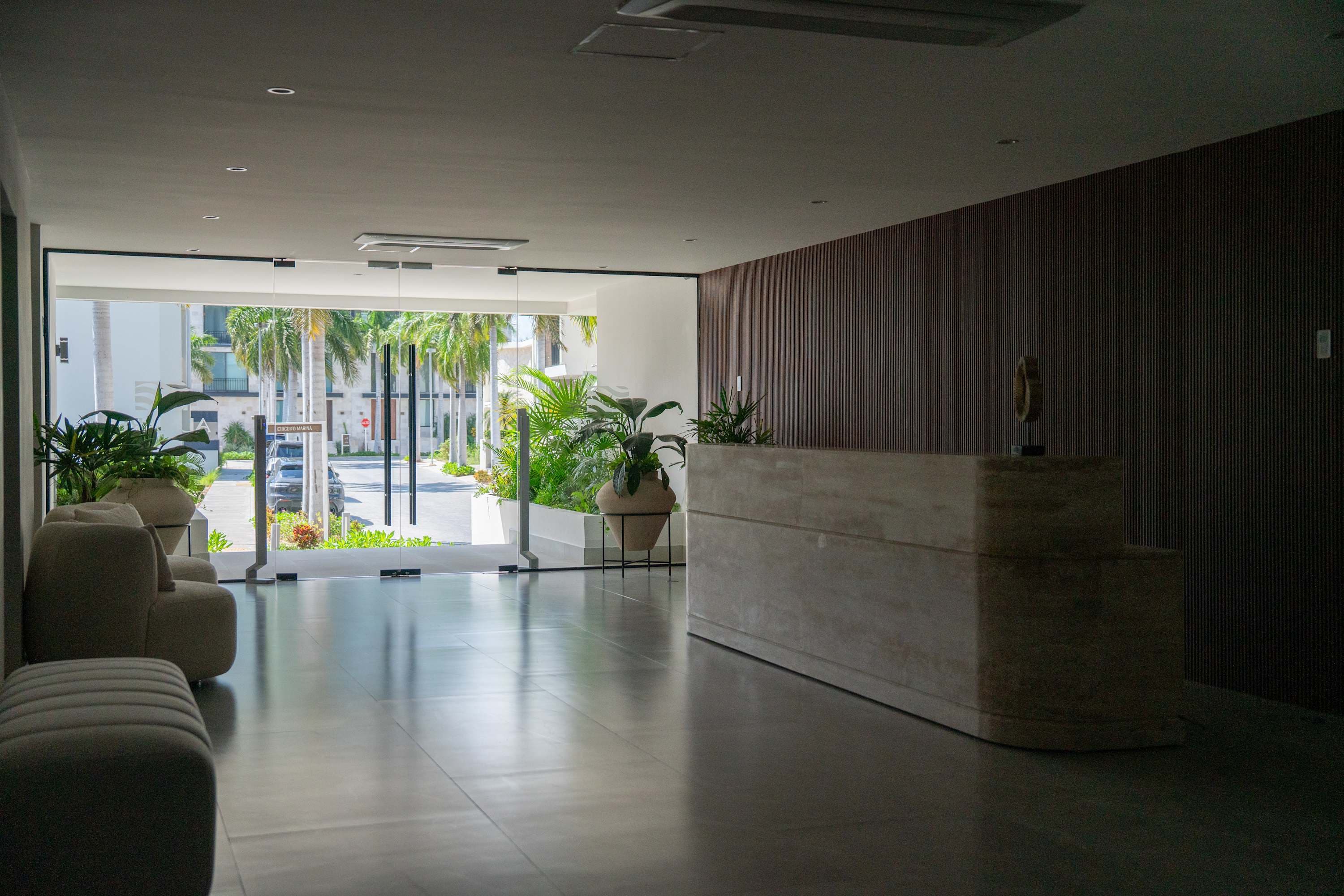 Modern lobby with reception desk, seating, and plants, overlooking a street with palm trees.