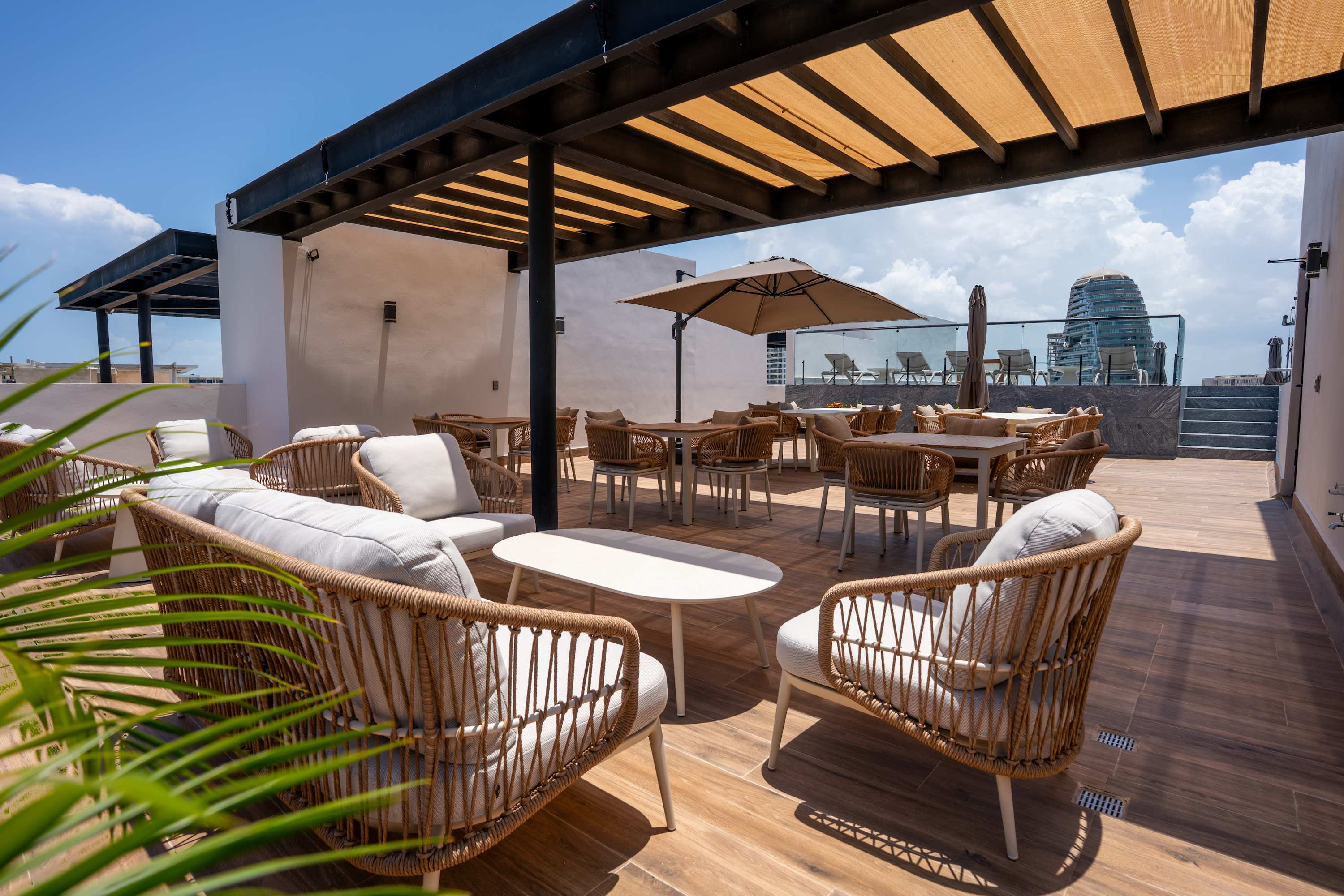 Rooftop patio with wicker furniture, tables, chairs, and umbrellas, overlooking a modern building.
