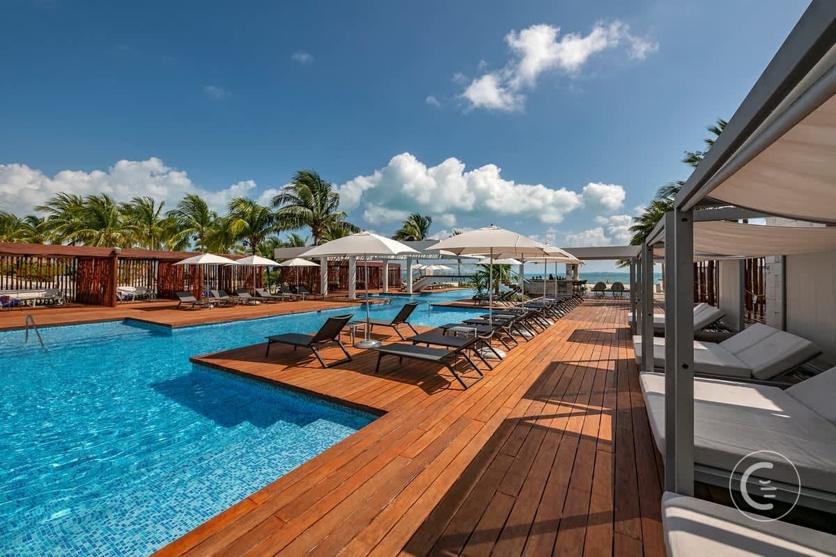 Resort pool with lounge chairs, cabanas, umbrellas, palm trees, and the ocean in the background.