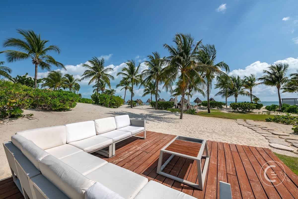 White sectional sofa and coffee table on a wooden deck overlooking a sandy beach with palm trees.