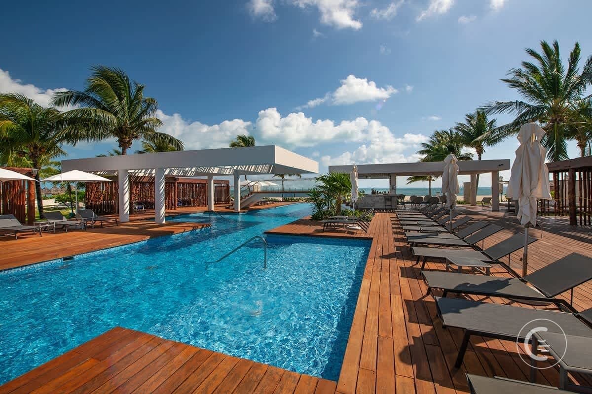 Resort pool with lounge chairs and palm trees under a cloudy blue sky.
