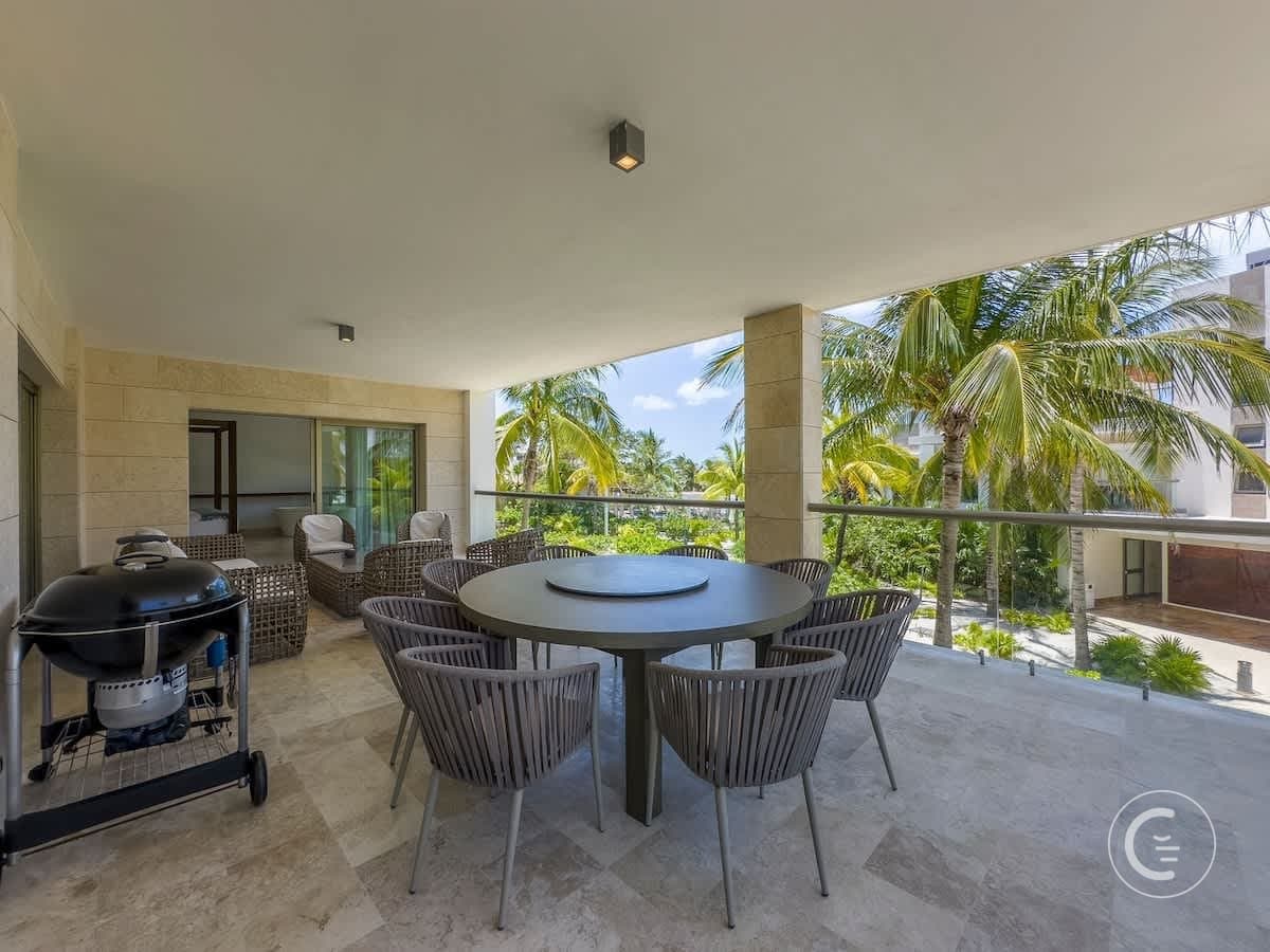A balcony with a dining table, chairs, and a grill overlooks palm trees.