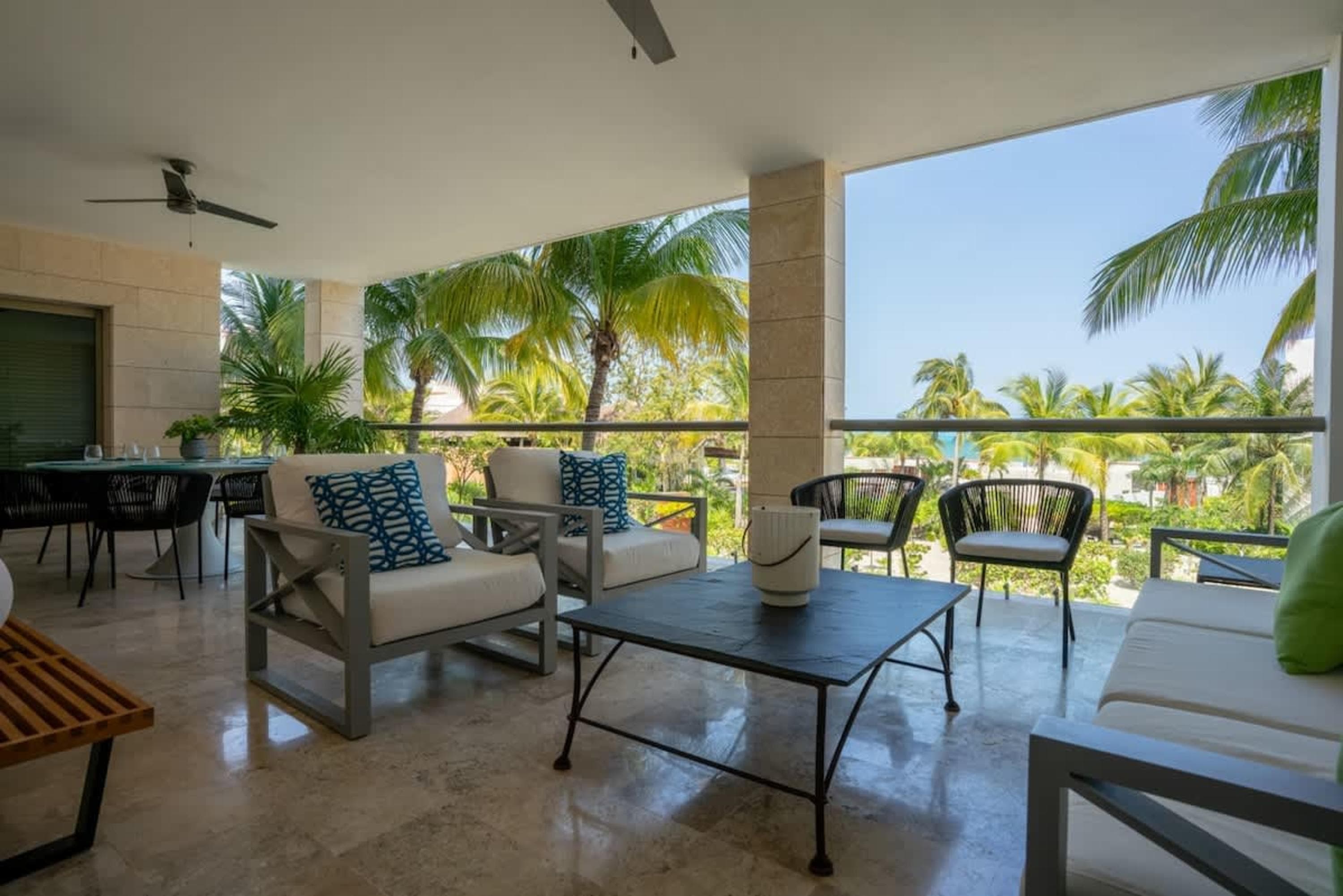 A balcony with chairs, sofas, and a table overlooking palm trees and the ocean.