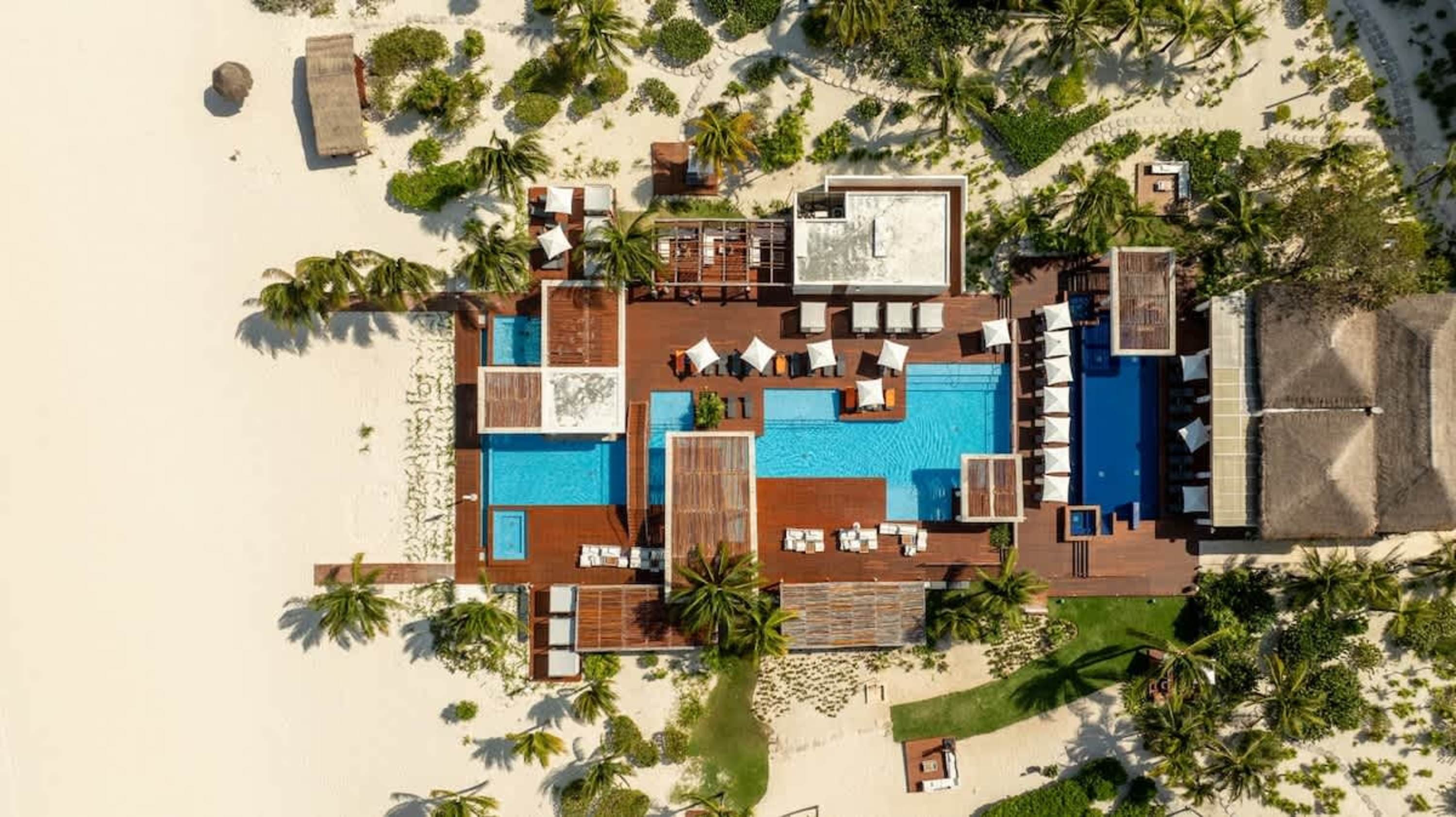 Aerial view of a resort with pools, lounge chairs, and palm trees on a sandy beach.