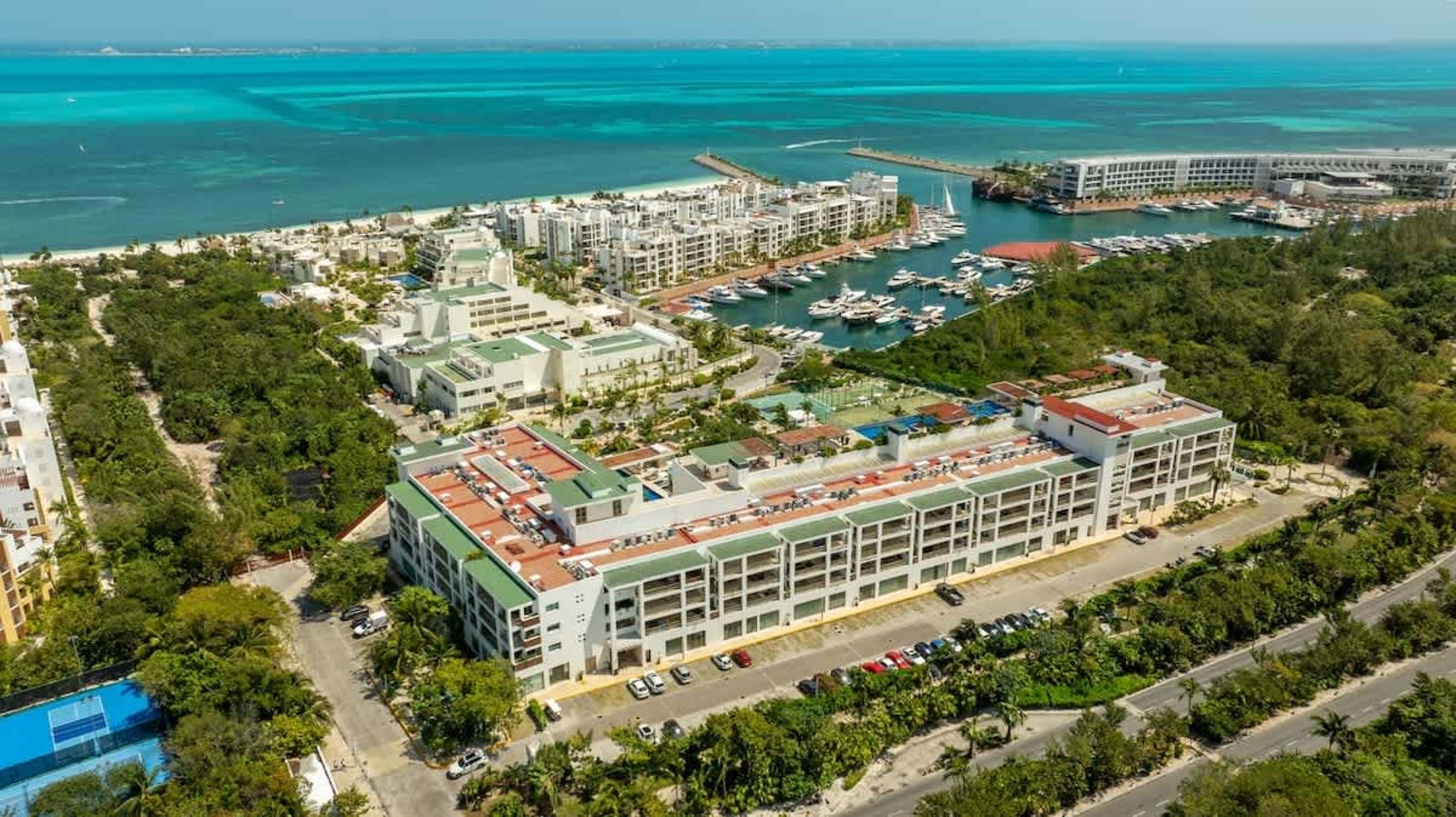 Aerial view of a modern resort complex with pools, tennis courts, and a marina on the coast.