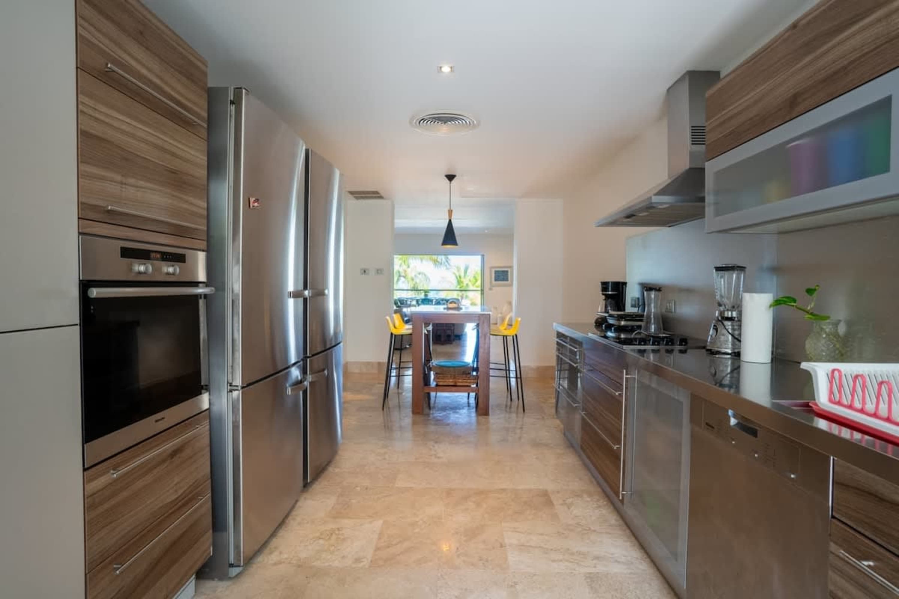 A modern stainless steel kitchen with an oven, refrigerator, and a dining area with yellow stools.