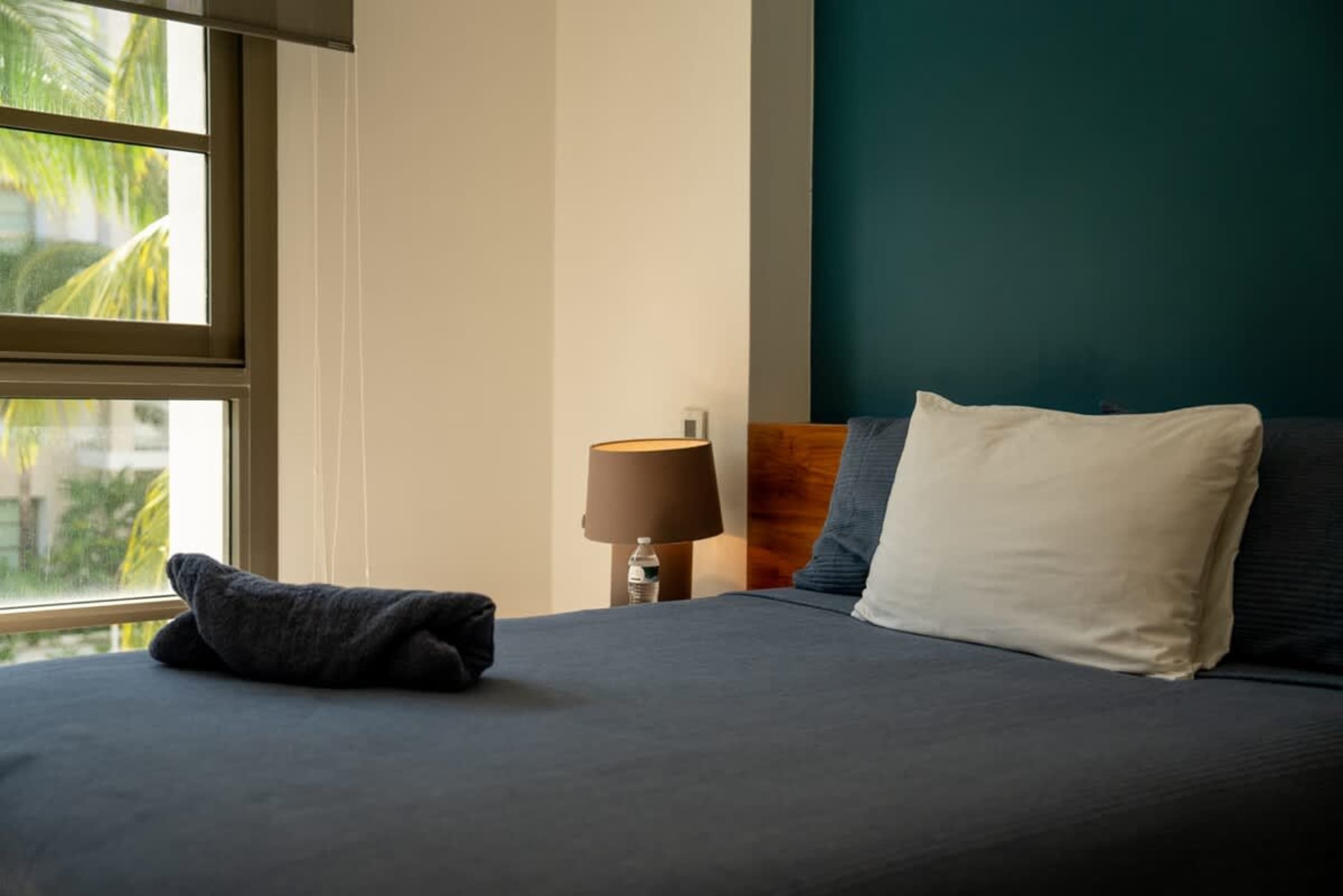 A bed with a blue comforter, white pillow, and a dark blue towel rolled up.