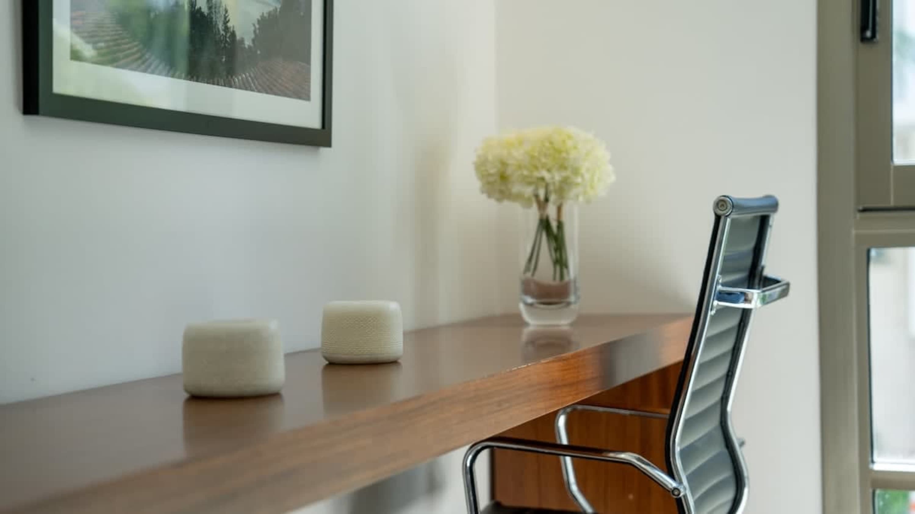 A wooden desk with two decorative white candles, a vase of white flowers, and a black office chair.