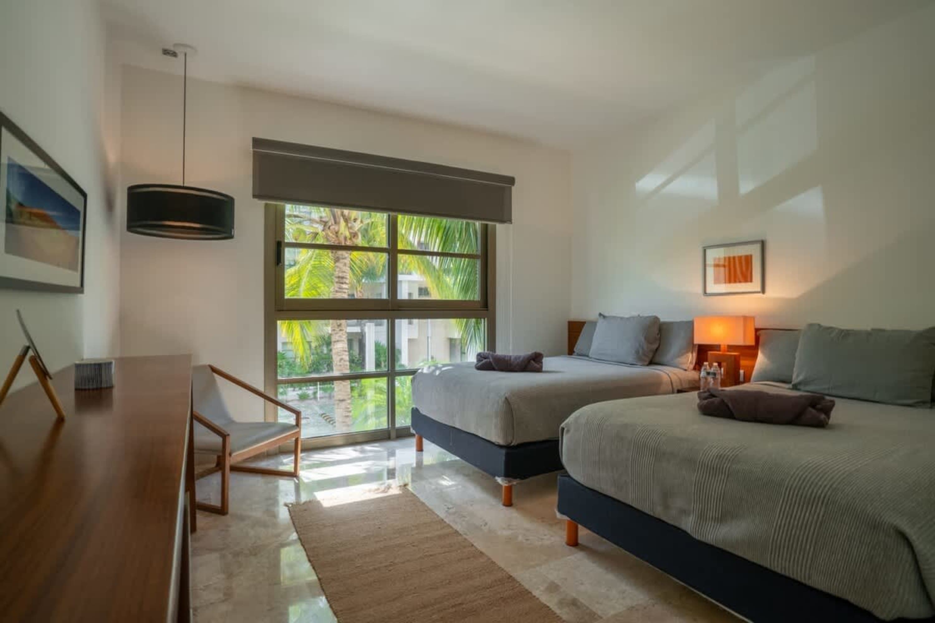 A bedroom with two beds, a window overlooking palm trees, and a wooden dresser.