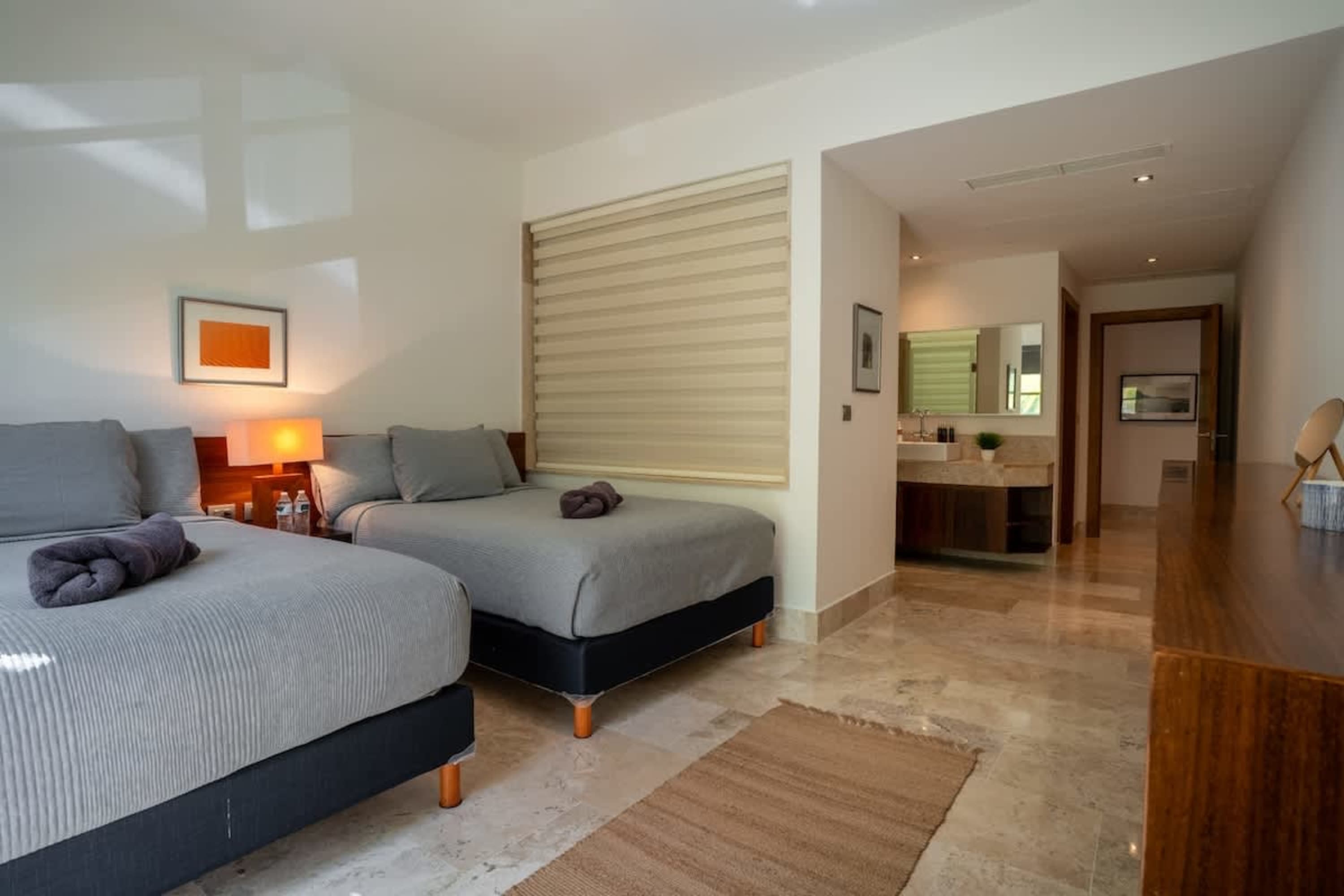 Two beds with grey linens are in a bedroom. A dresser and doorway are visible in the background.