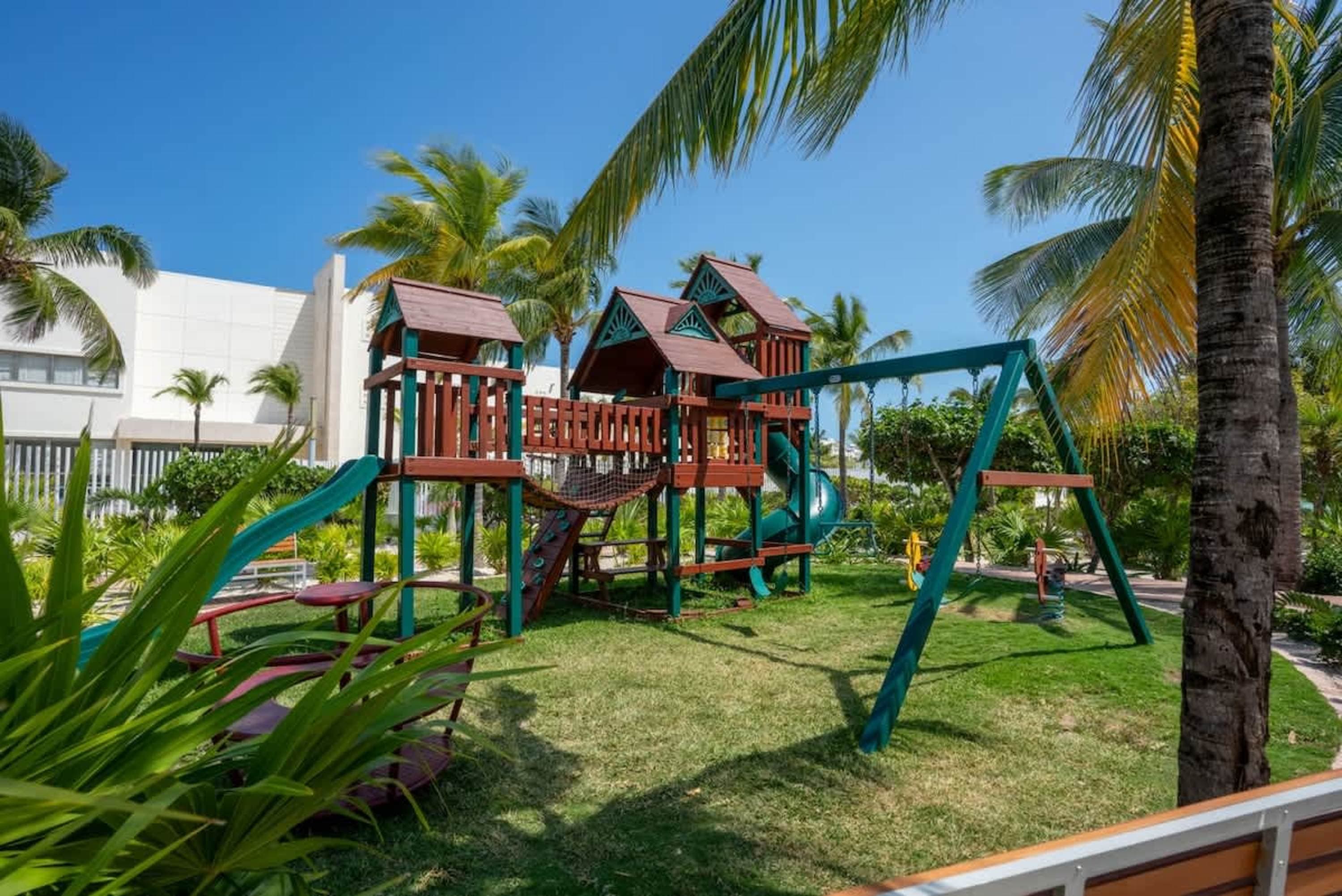 Playground with slides, swings, and climbing nets under palm trees with a white building in the back