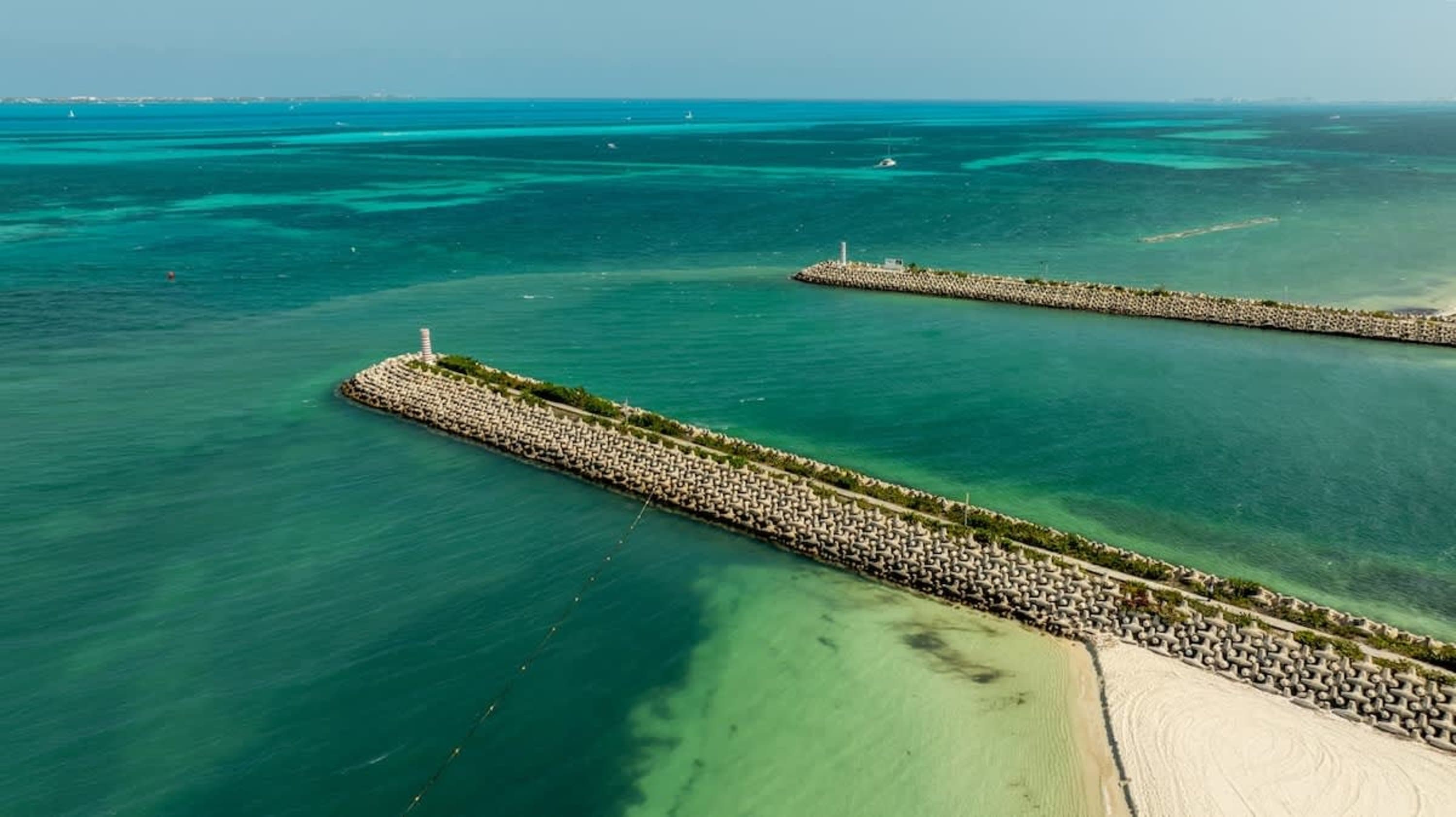 Aerial view of breakwaters in turquoise water, with a beach and a lighthouse.