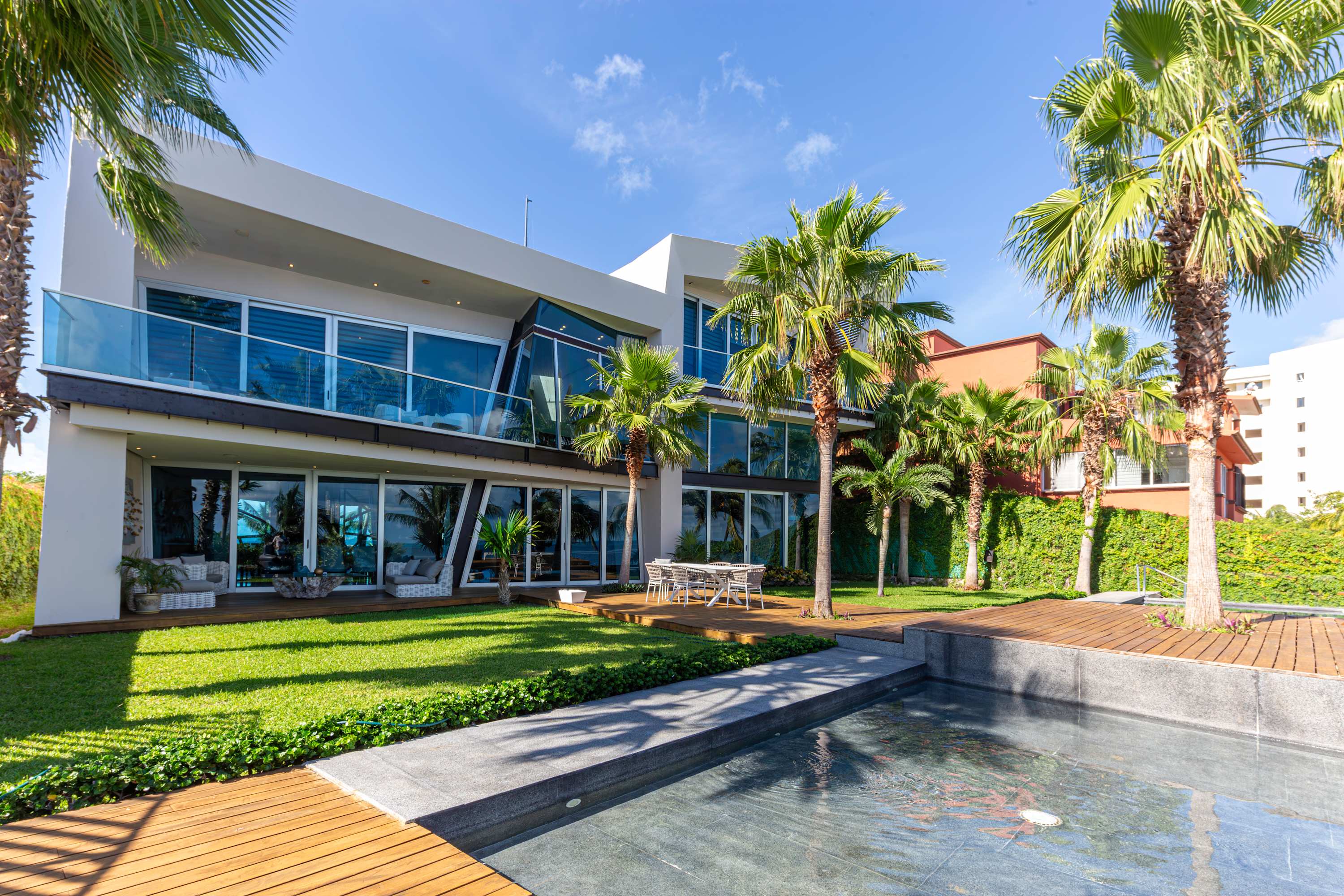 Modern house with glass walls and balconies overlooking a lush green lawn and pool.