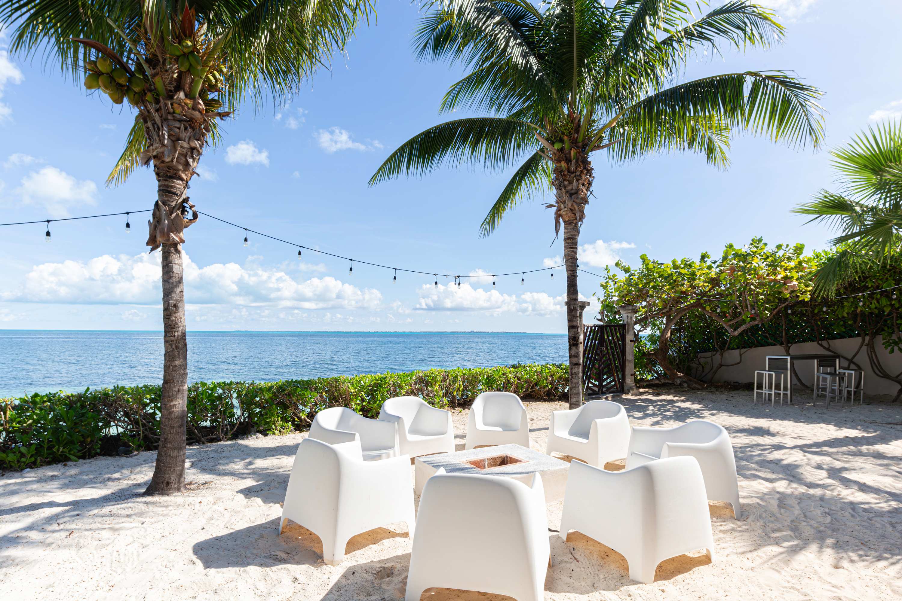 A circular seating area with white chairs surrounds a fire pit on a sandy beach.