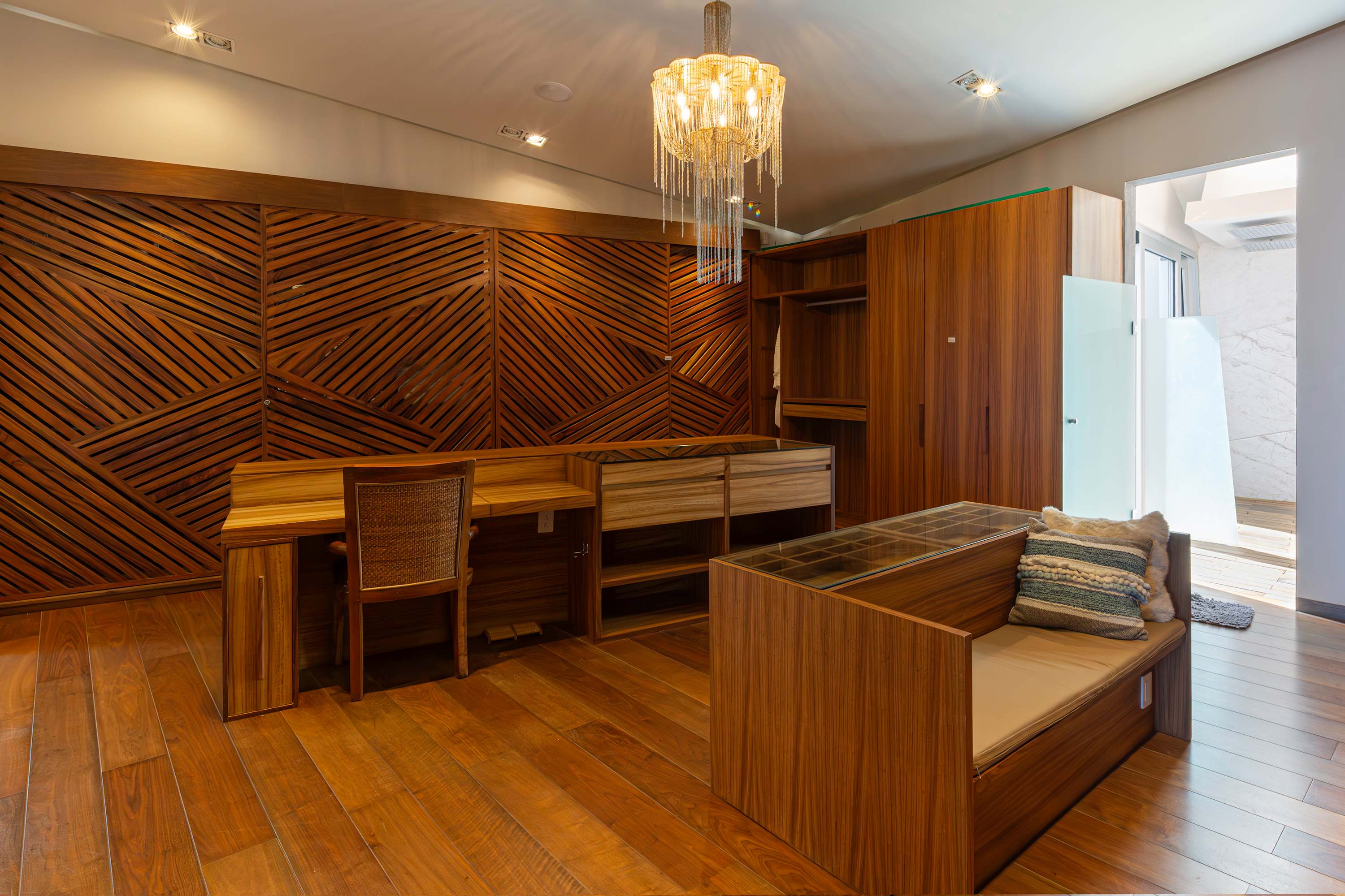 Luxurious wooden walk-in closet with a desk, chandelier, and a bench with pillows.