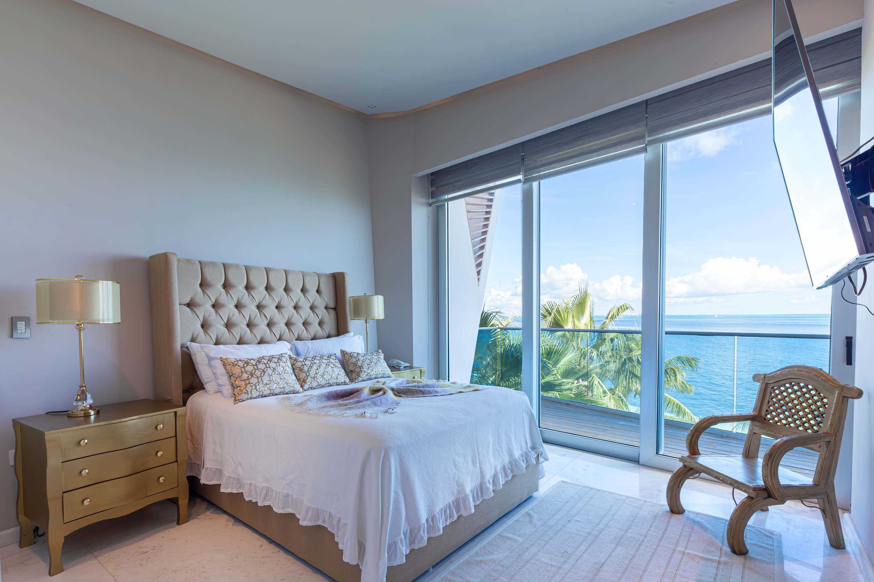 A bedroom with a large bed, a nightstand, and a chair, facing a balcony with an ocean view.