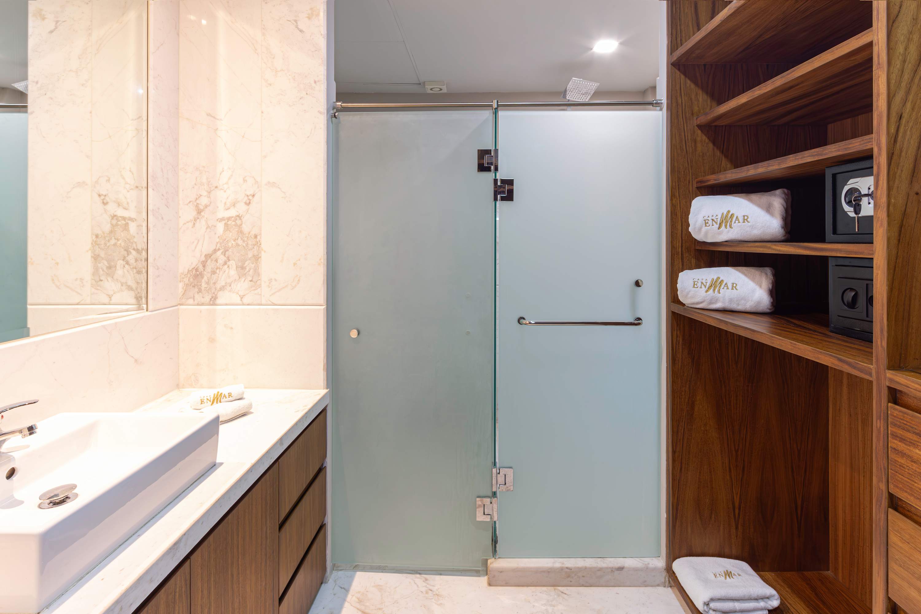 A modern bathroom with a marble vanity, frosted glass shower, and wooden shelves with safes and towe
