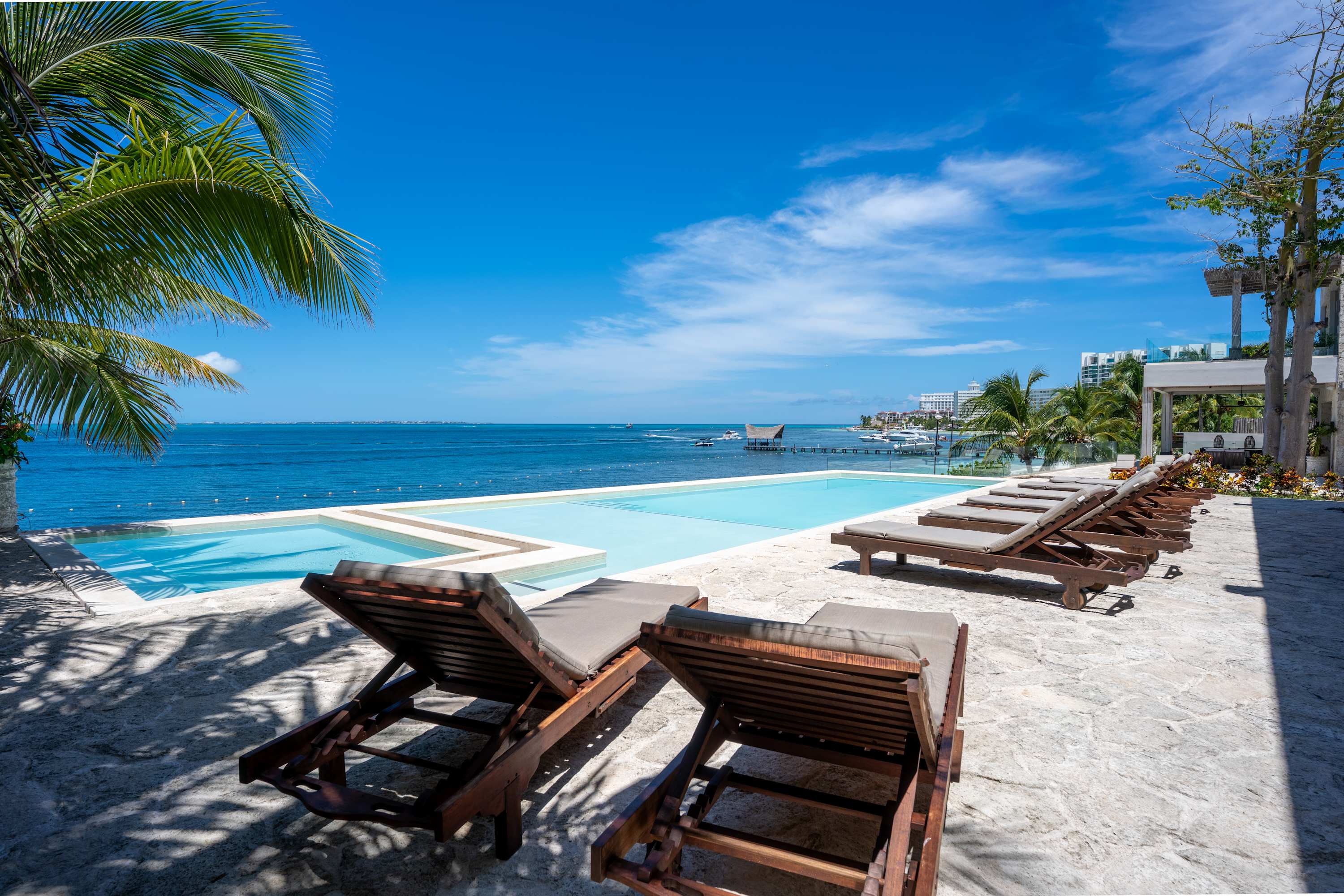 Lounge chairs near a pool overlook the ocean with boats.