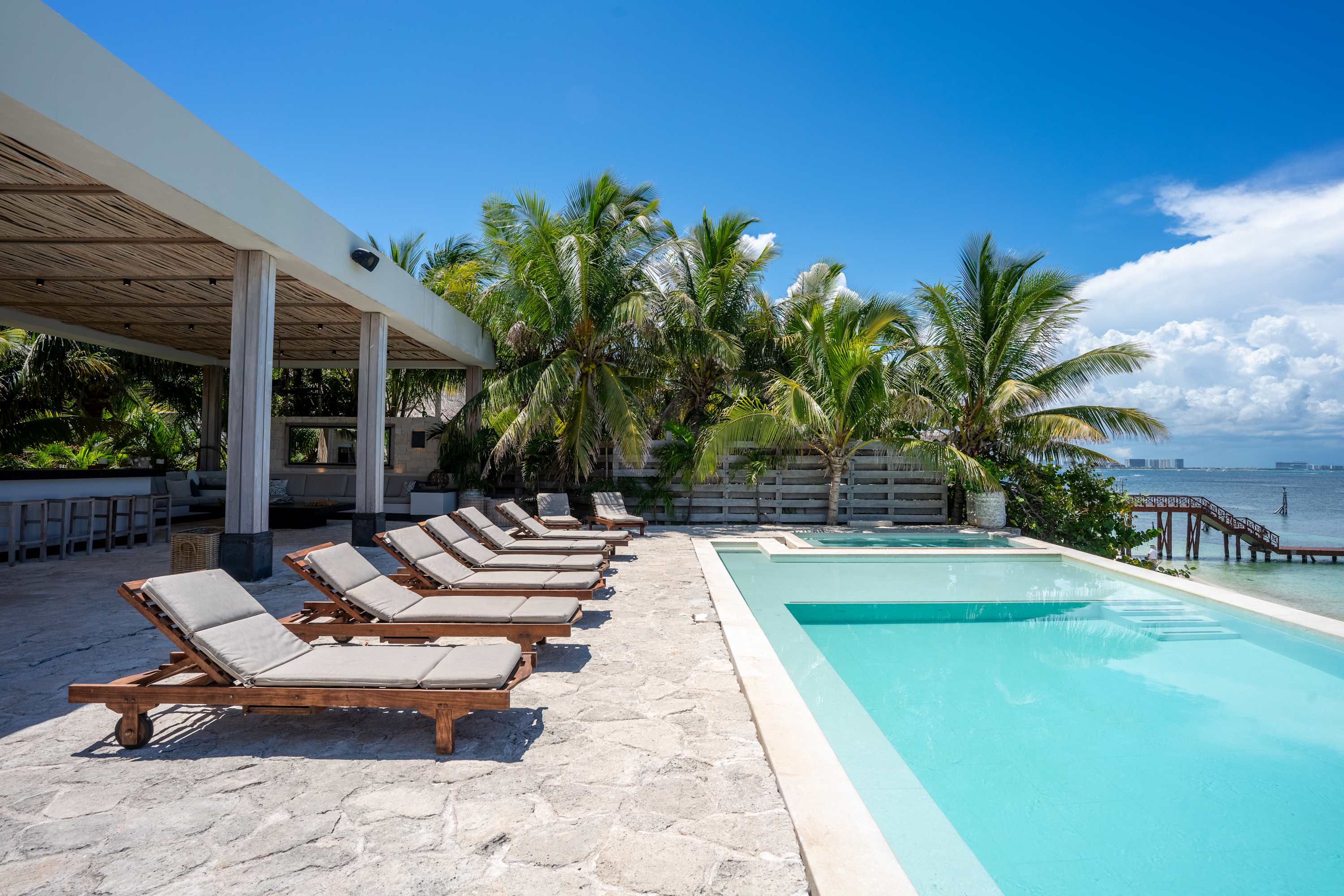 Lounge chairs line a stone patio next to a bright blue pool, with palm trees and the ocean in the ba