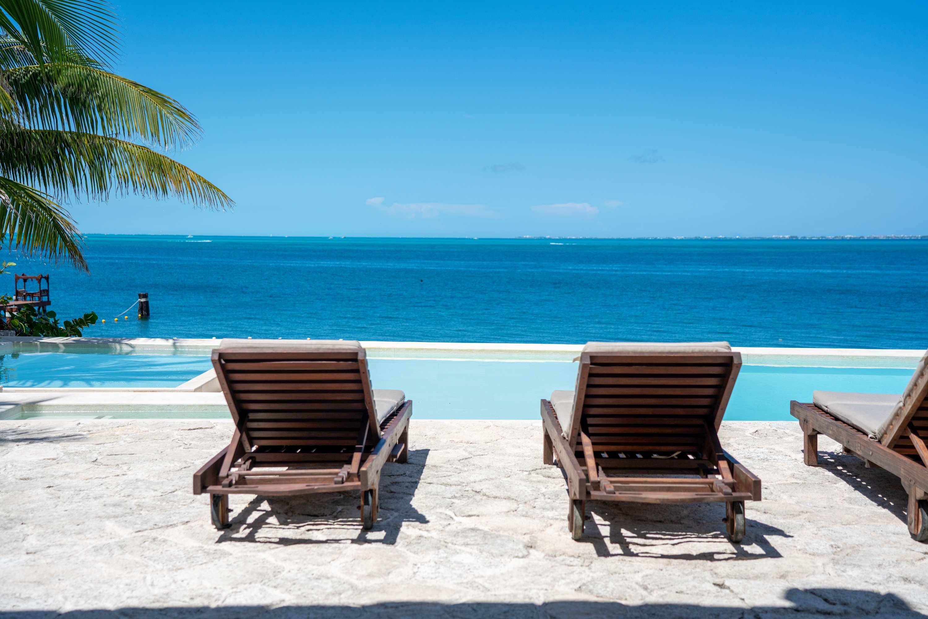 Deck chairs face a tranquil infinity pool and the azure ocean on a sunny day.