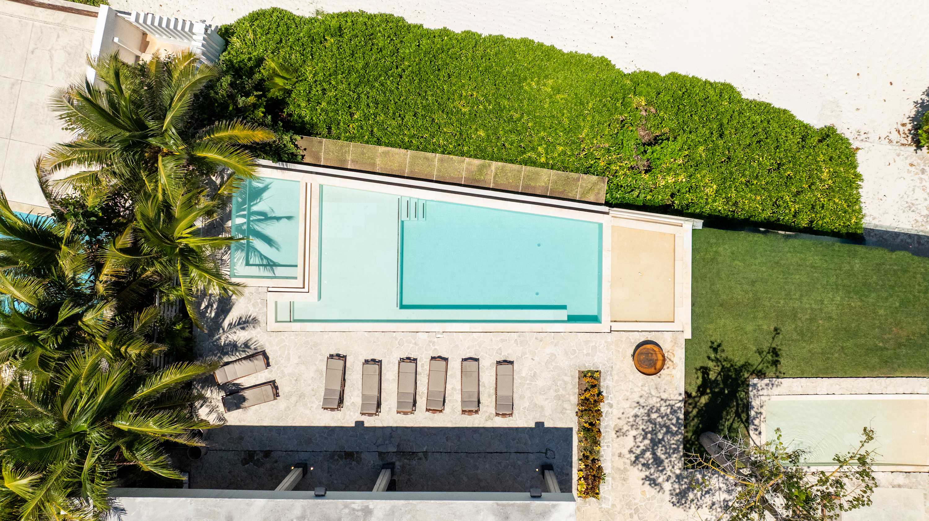 An aerial view of a modern villa with a pool, lounge chairs, palm trees, and lush greenery.