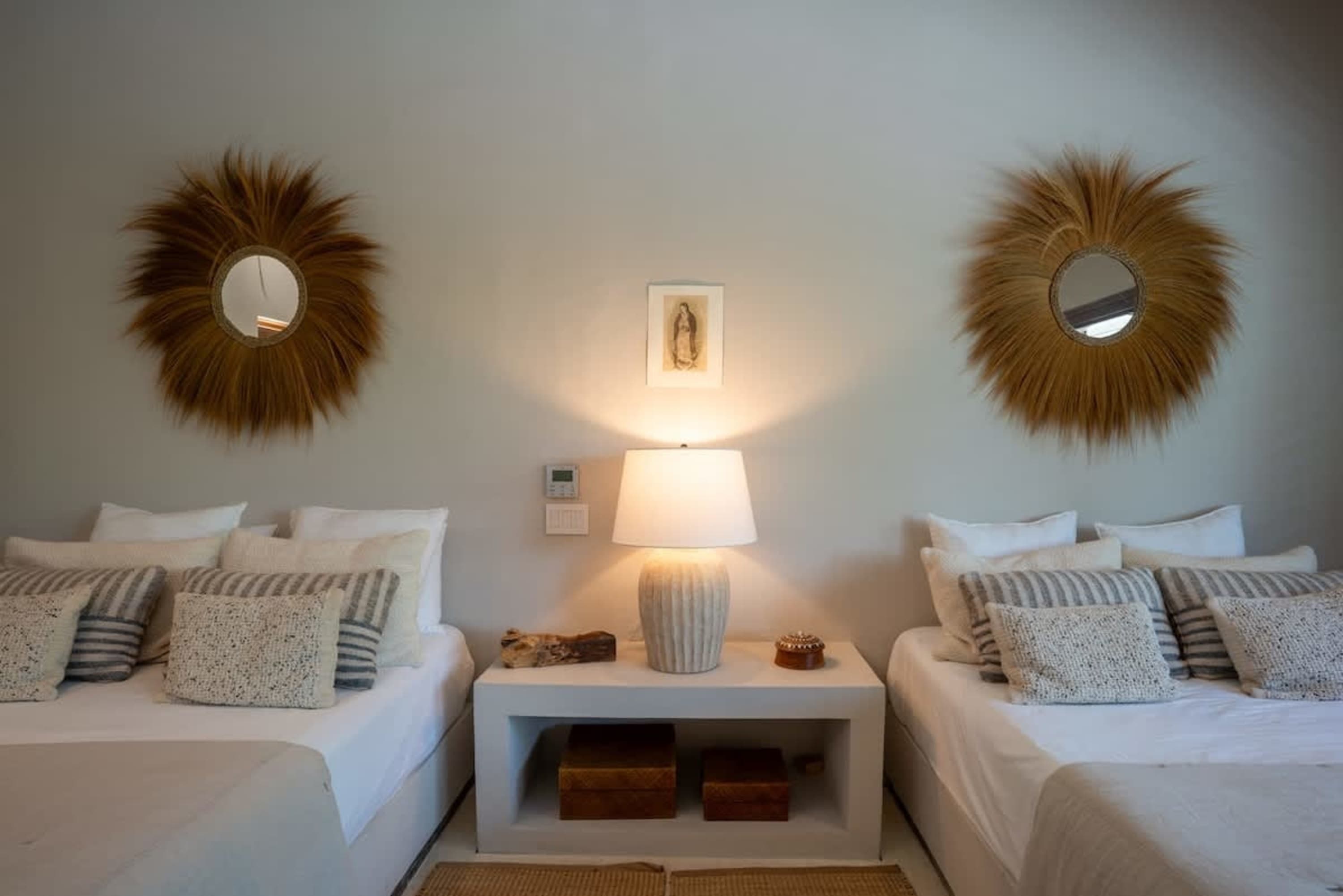 A serene bedroom with two beds, fluffy pillows, and a decorative sunburst mirror above each bedside.