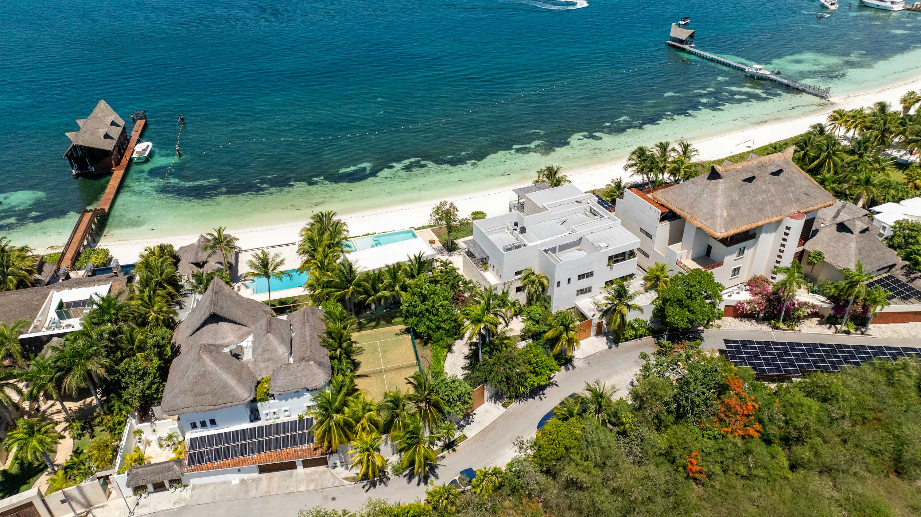 An aerial view of a tropical resort with thatched-roof villas, a tennis court, swimming pools, and a