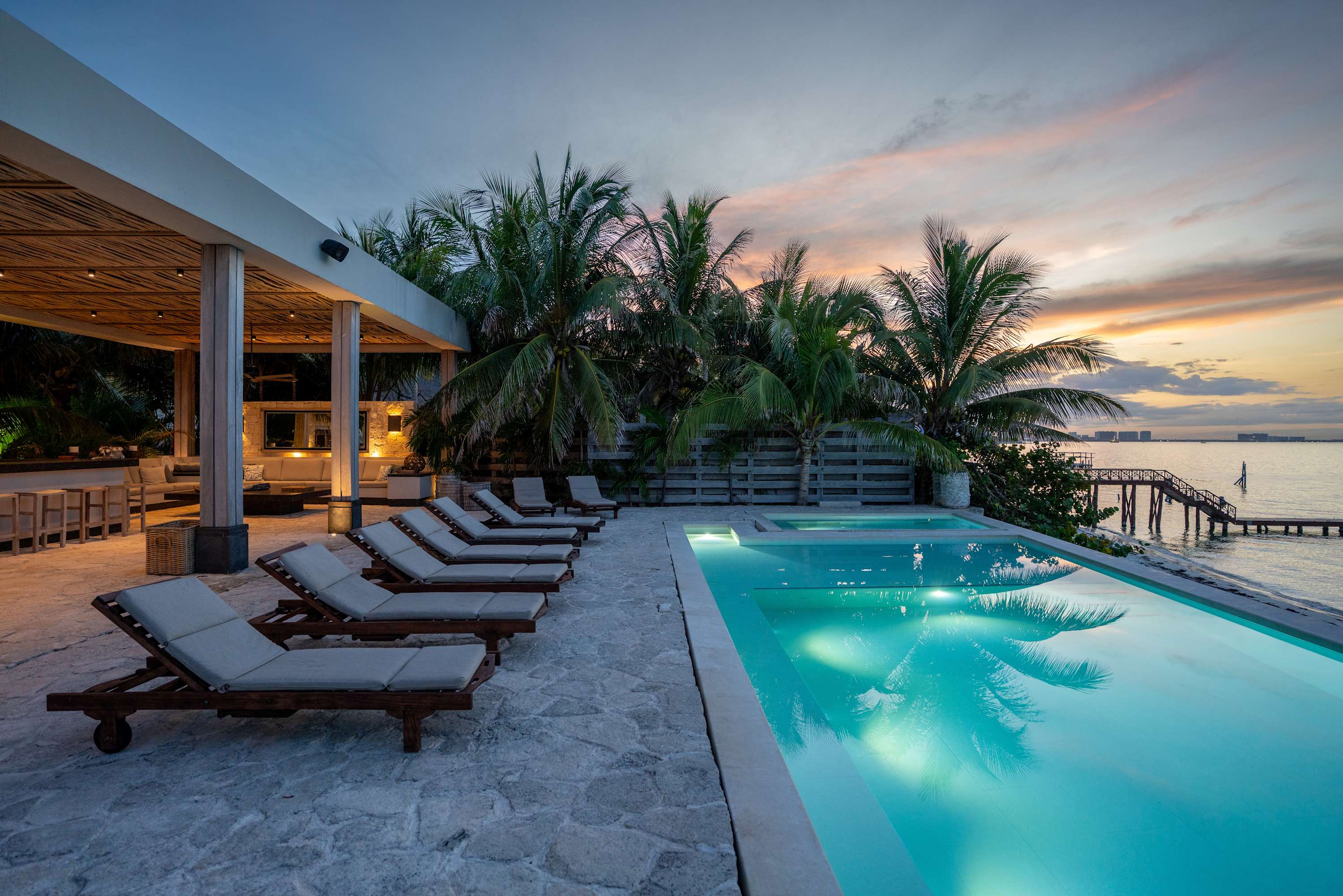 Infinity pool and loungers at sunset with palm trees and a view of the ocean.