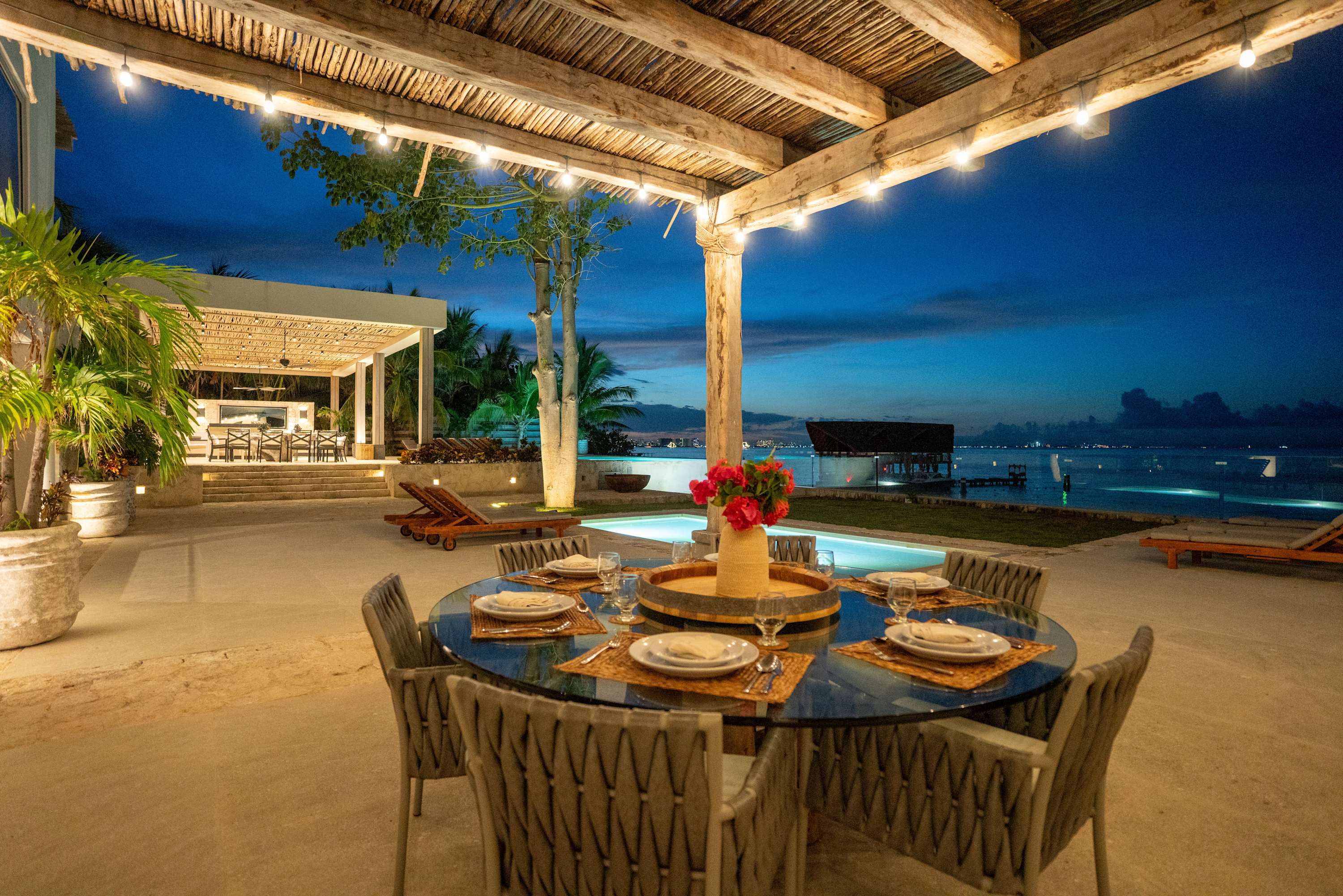 An outdoor dining area with a glass-top table set for dinner overlooks a tranquil ocean at dusk.