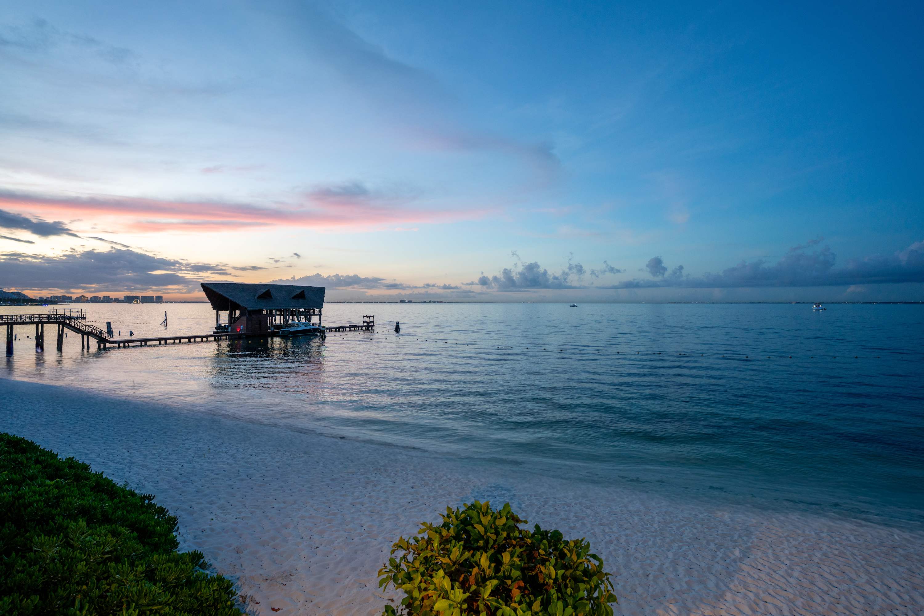 A tropical sunrise with pink and orange clouds over a calm ocean, a pier, and a thatched-roof buildi