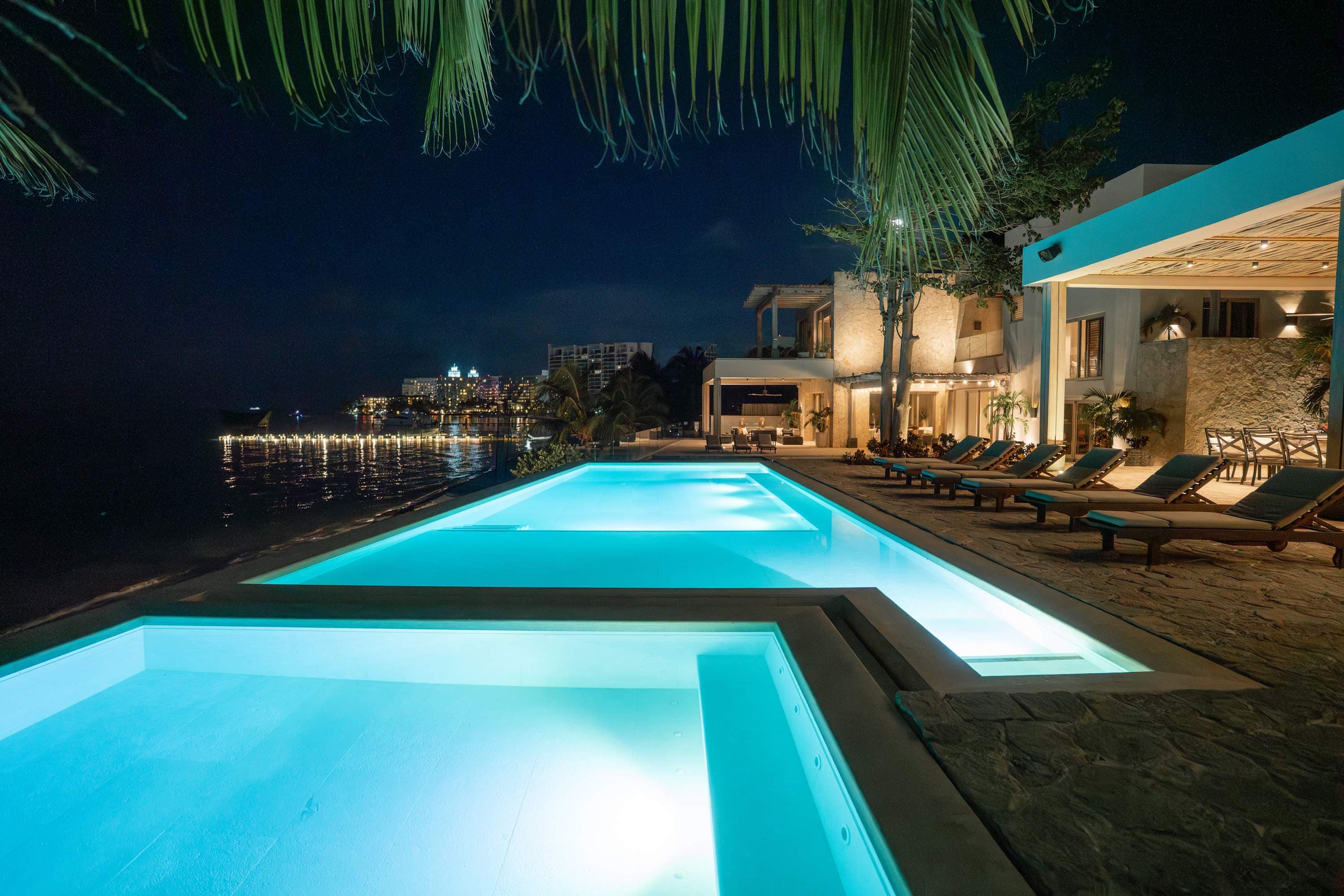 A luxurious infinity pool glows blue at night beside a tropical resort and calm ocean.