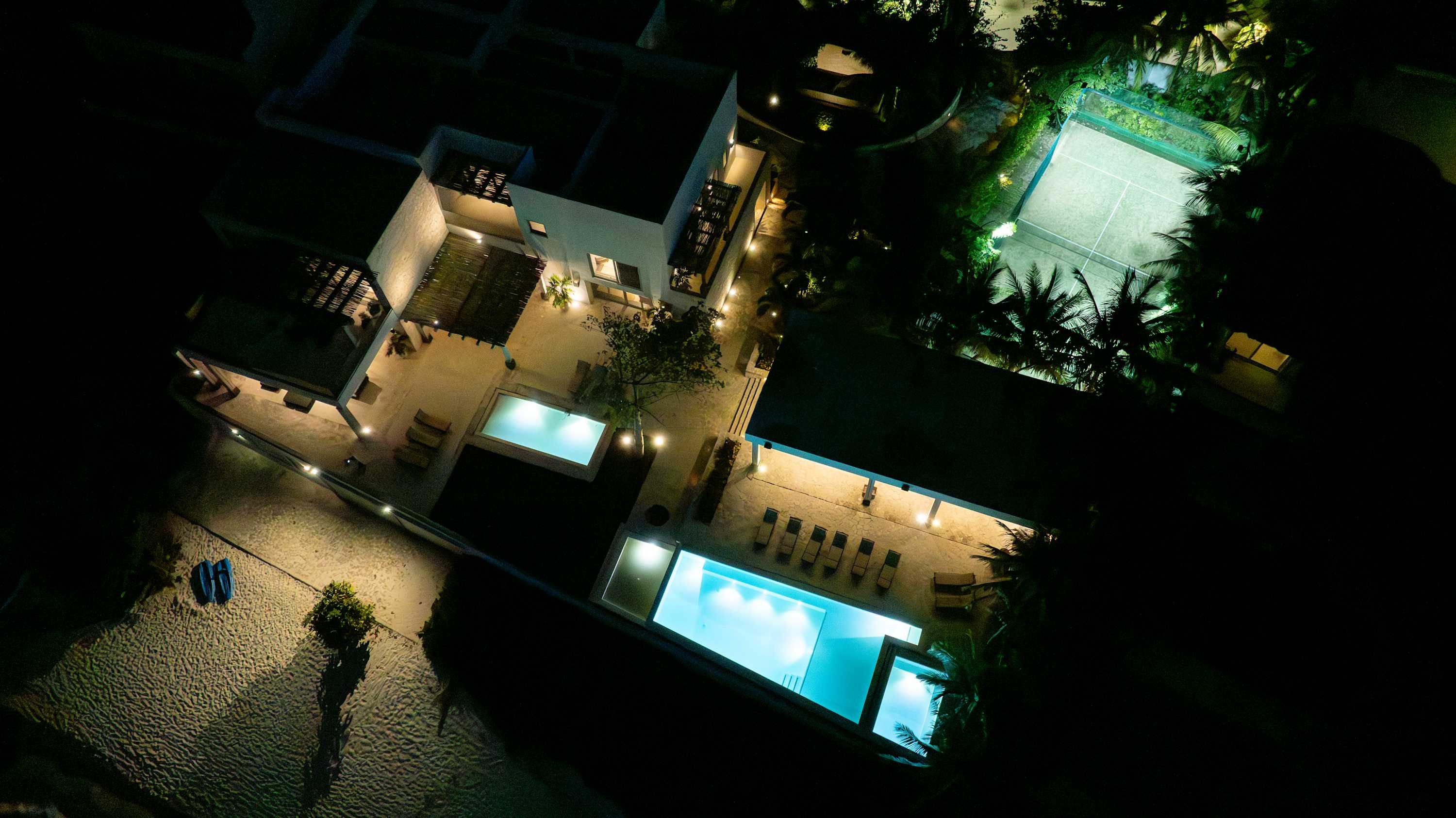 Aerial night view of a resort complex with pools, a tennis court, and palm trees.