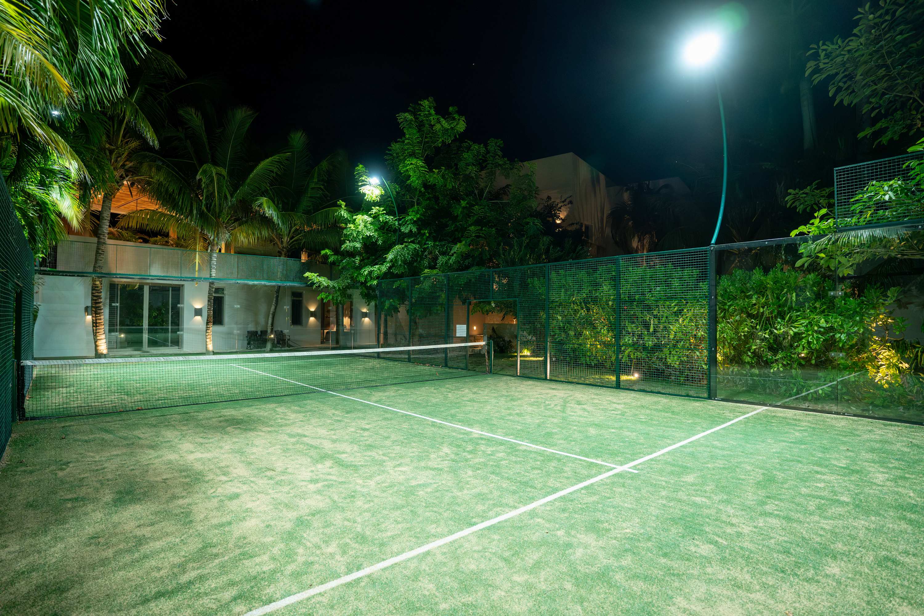 A night view of a brightly lit padel court surrounded by lush greenery and palm trees.