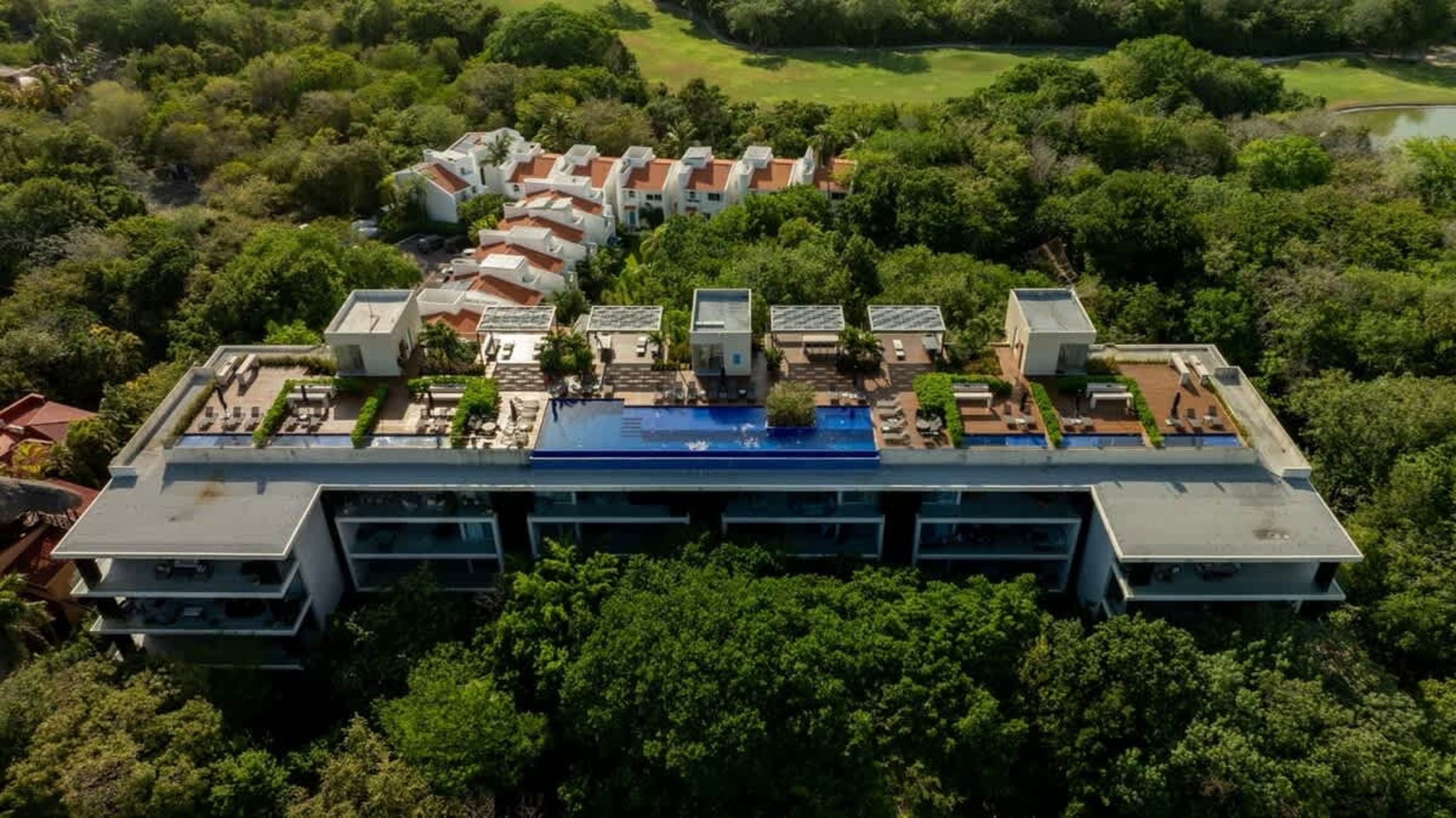Aerial view of a modern building with a rooftop pool, surrounded by lush trees.