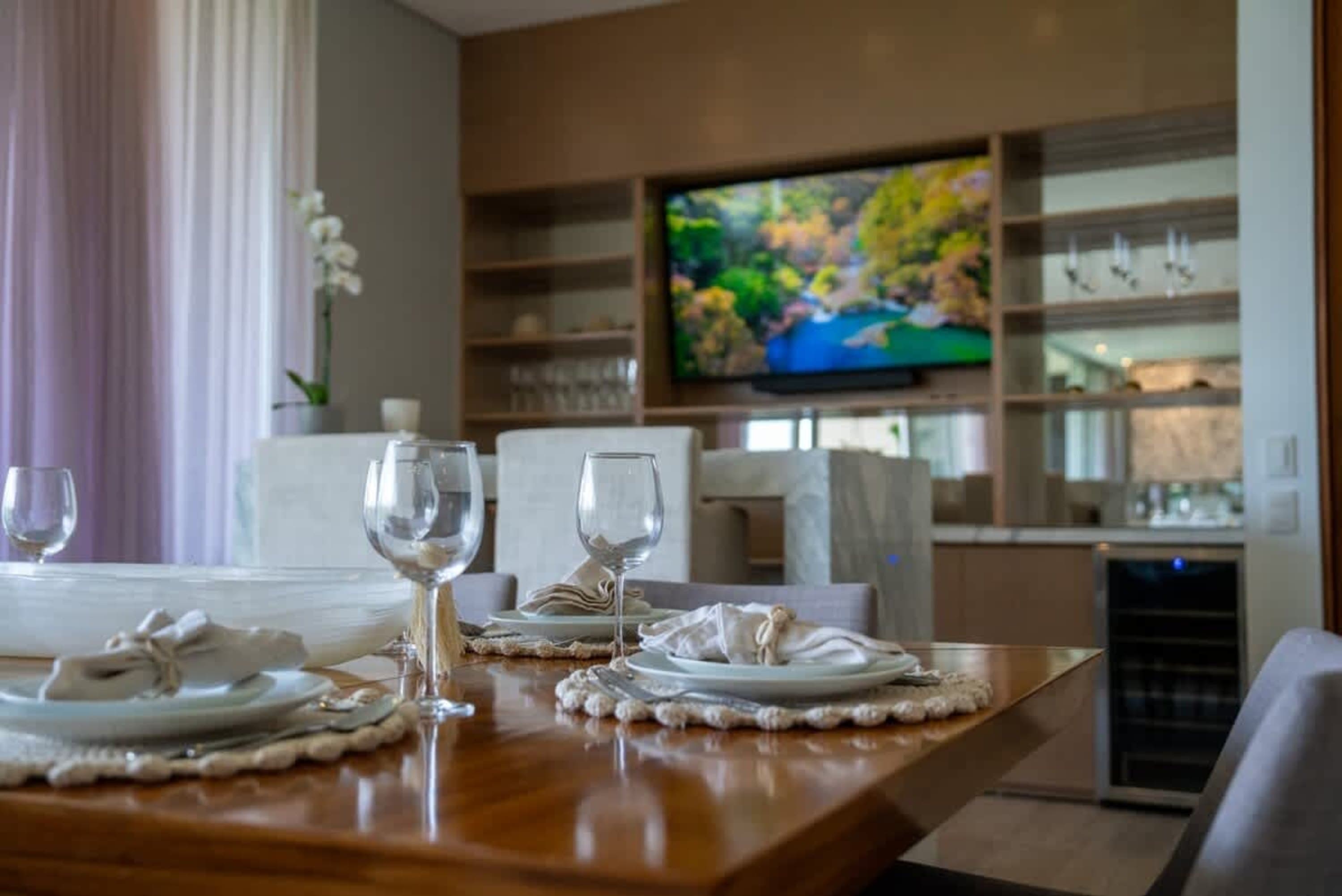 A dining table with place settings is in the foreground, with a TV and shelves in the background.