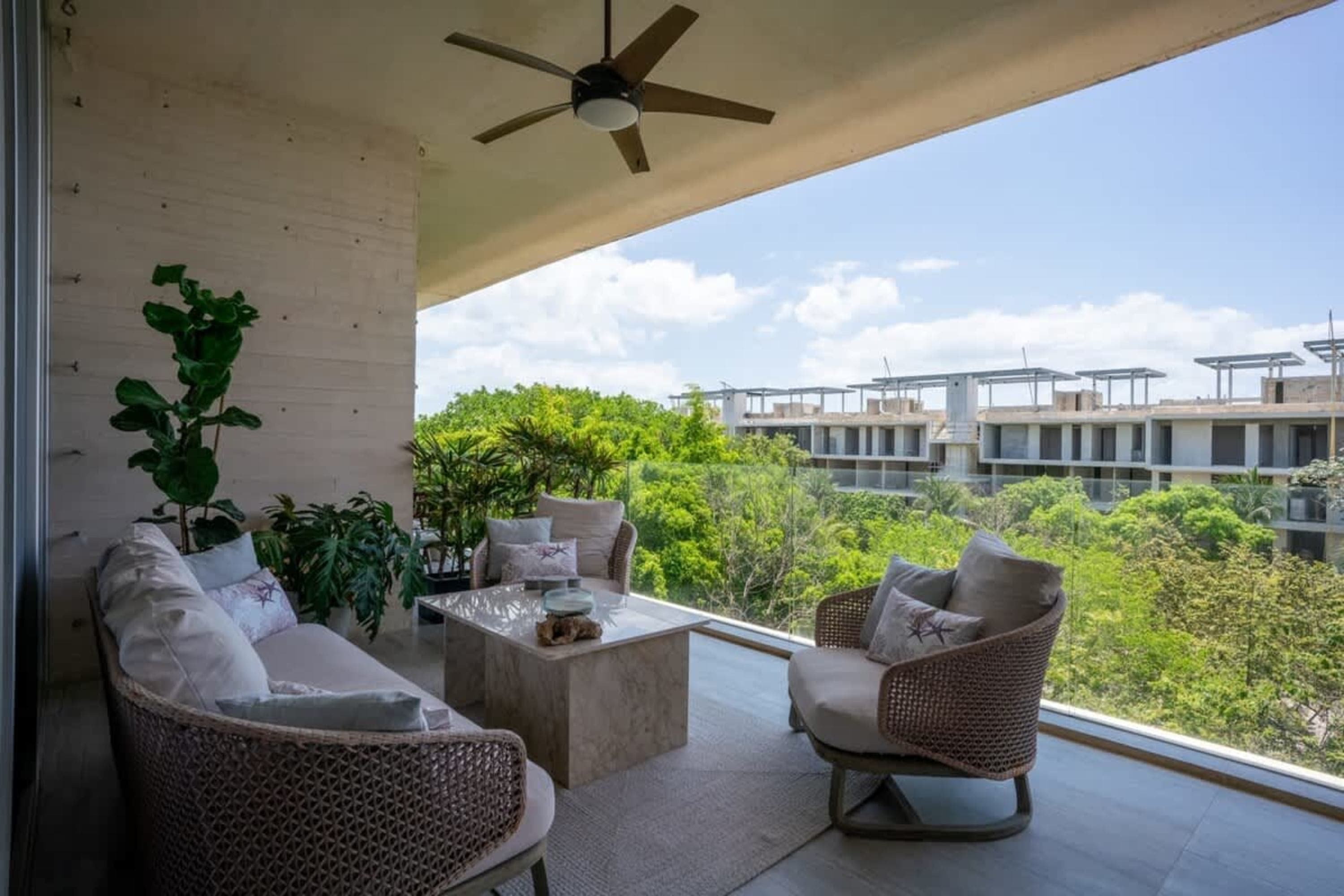 A covered balcony with rattan furniture and plants overlooks a lush green canopy and construction.