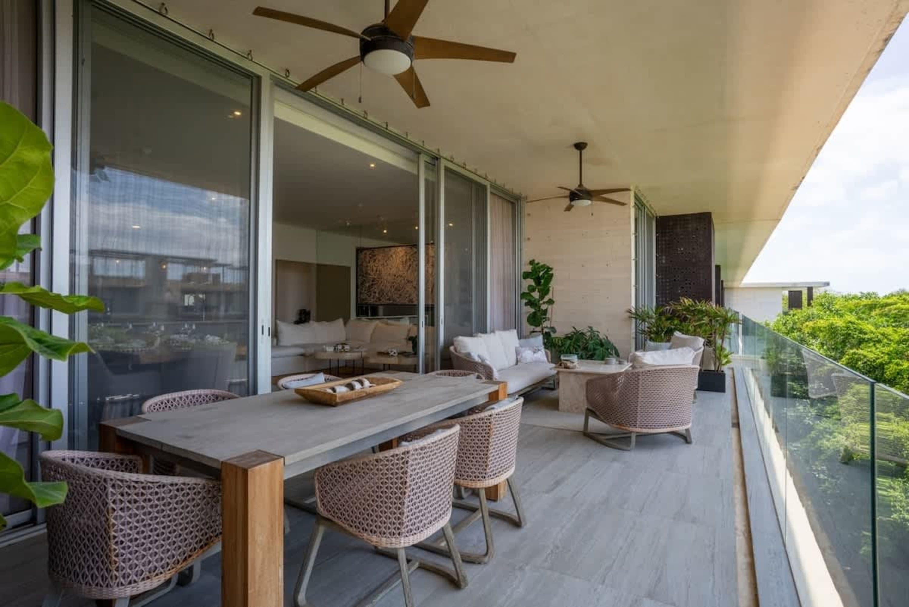 A modern balcony with a dining set, lounge seating, and glass railings overlooking lush trees.