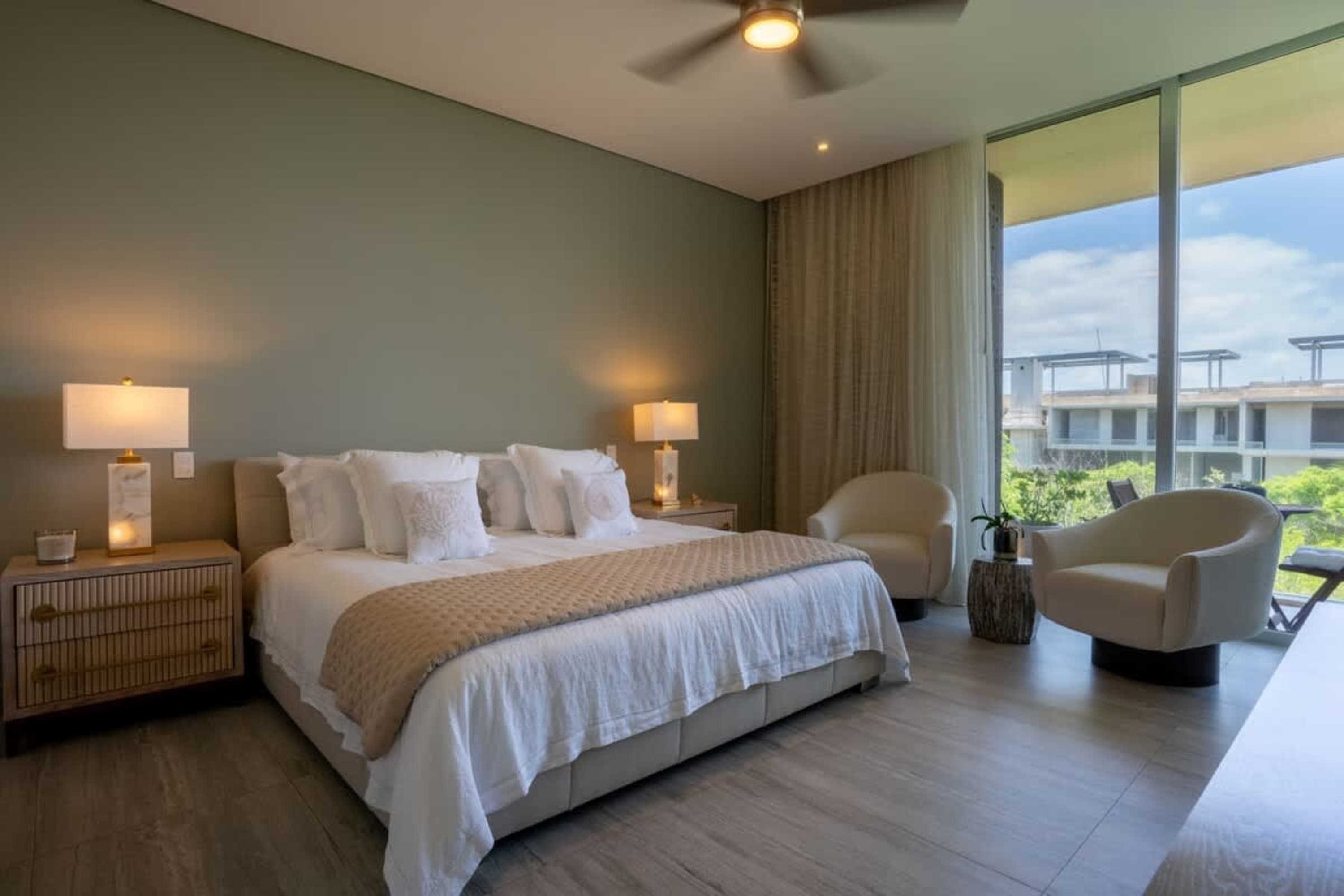A modern bedroom with a large bed, two chairs, and a scenic view from the balcony.
