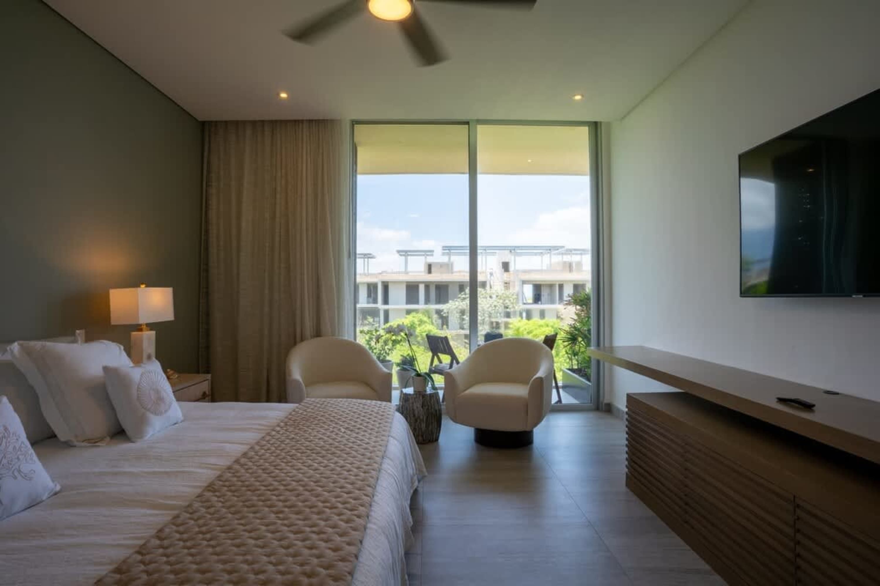 Modern bedroom with a bed, two chairs, and a TV overlooking a balcony with a city view.
