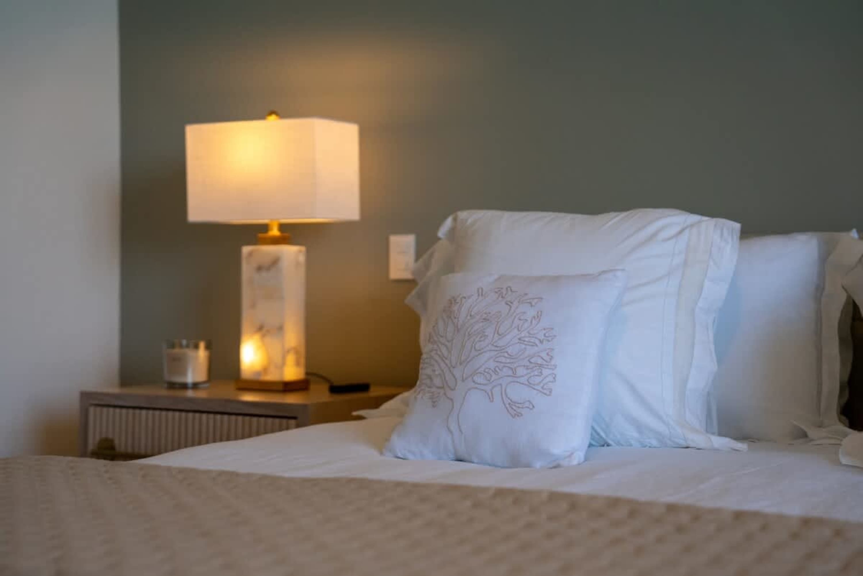 A close-up shot of a bed with white pillows and a taupe blanket, next to a nightstand with a lamp an