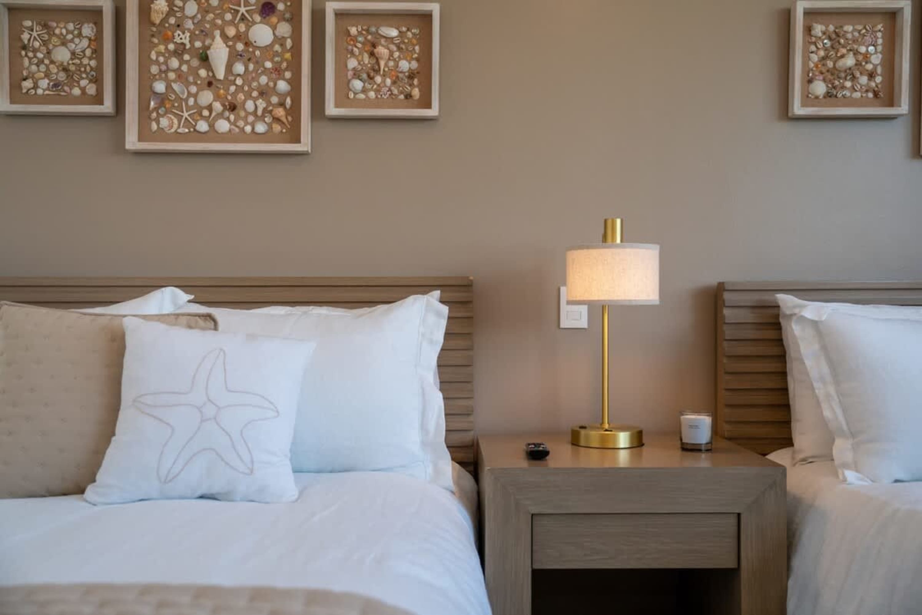Two beds in a room with a bedside lamp and seashell wall art.