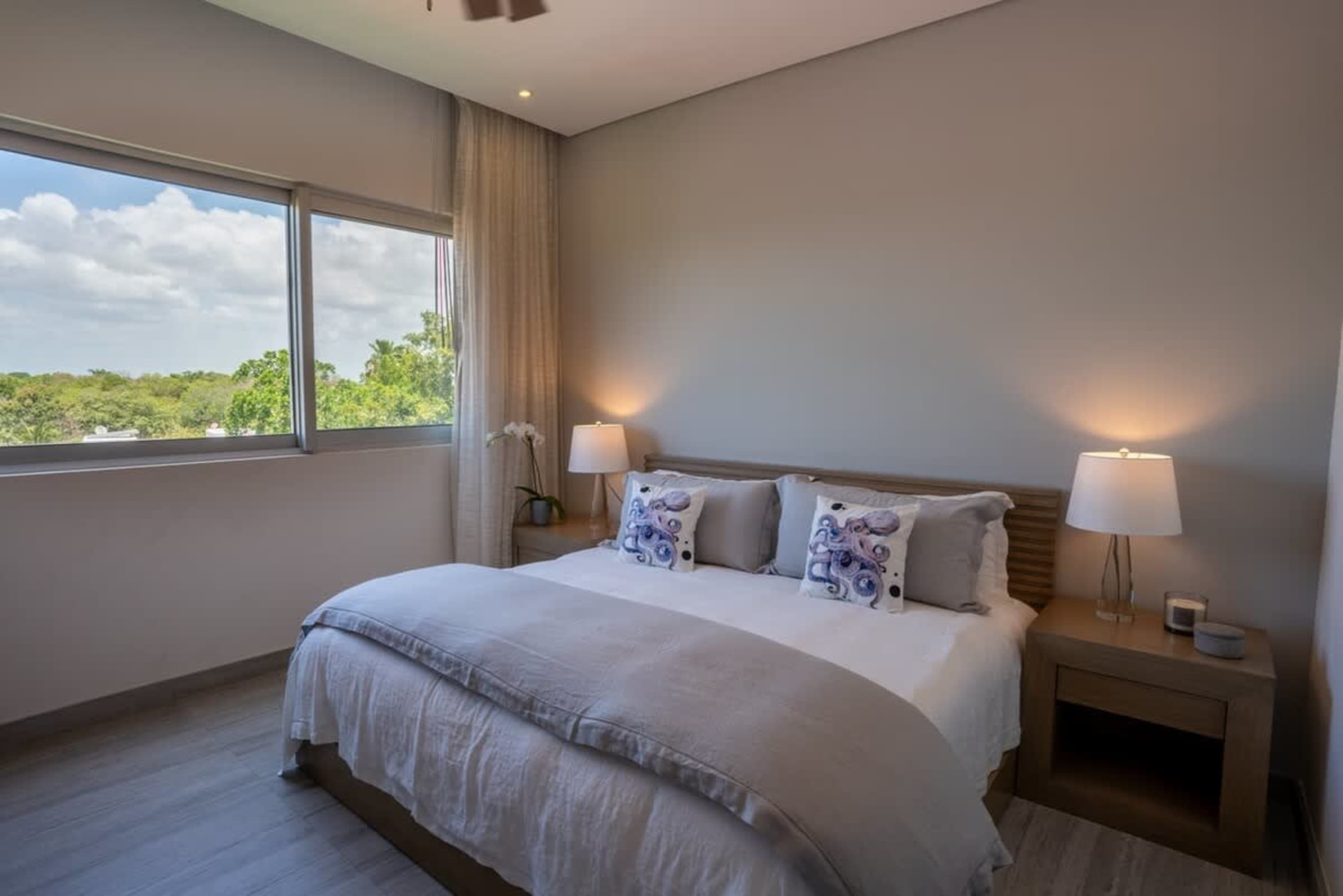 A modern bedroom with a large bed, octopus-themed pillows, and a view of lush greenery.