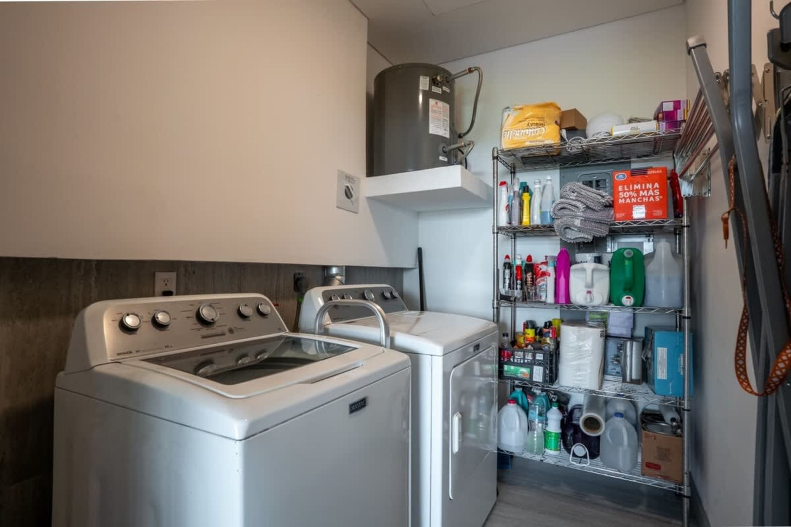 A laundry room with two white washers, a shelf of cleaning supplies, and a water heater.
