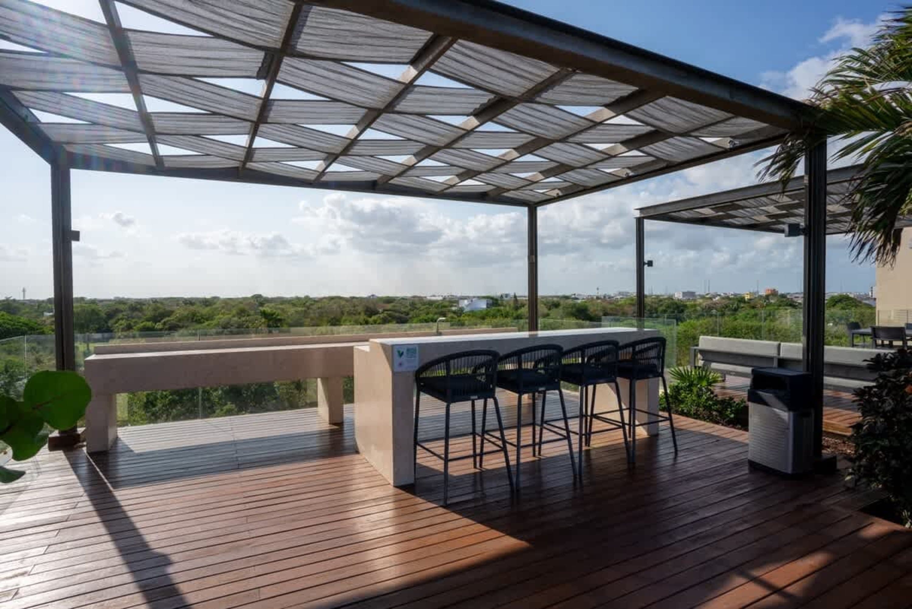 A modern outdoor bar with stools on a wooden deck, overlooking a lush green landscape under a pergol