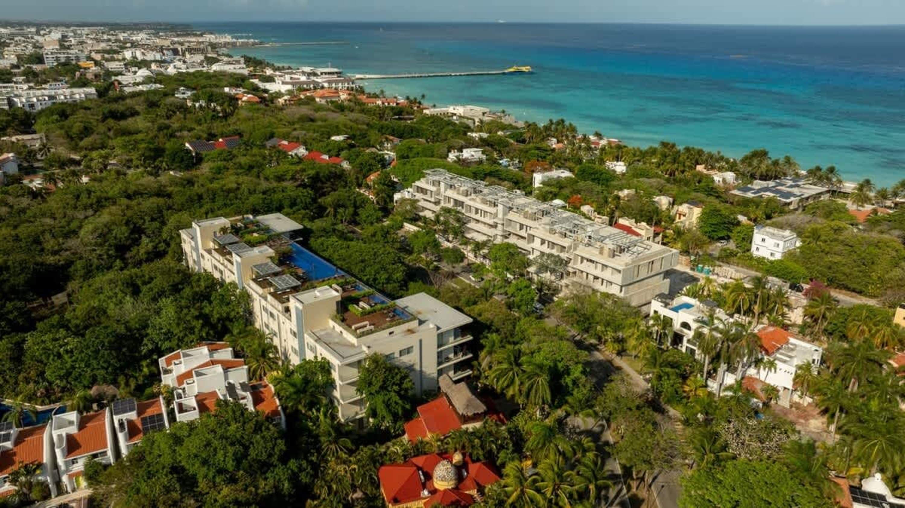Aerial view of a tropical resort town with modern buildings, lush green trees, and the blue ocean in